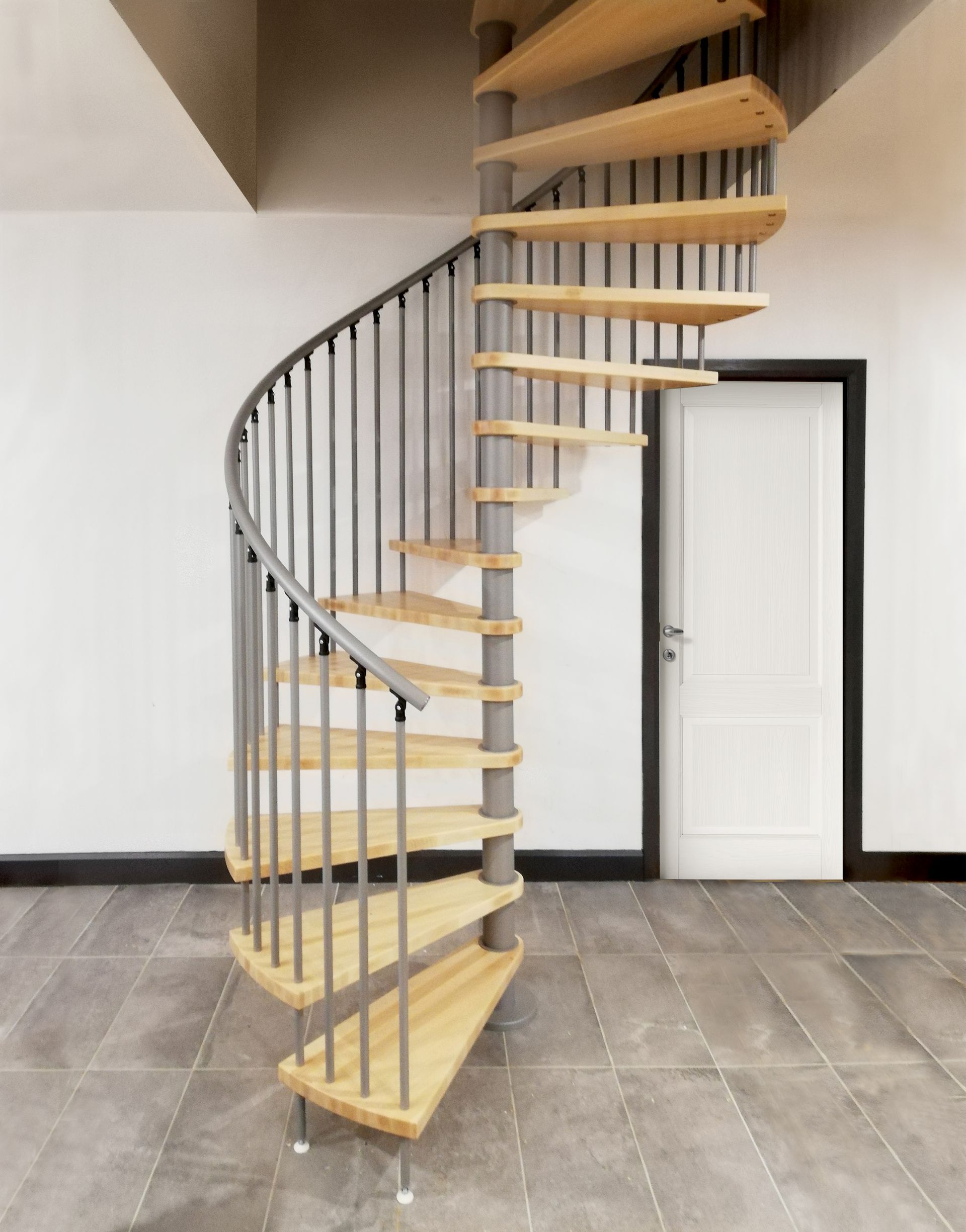 Scala a chiocciola con gradini in legno chiaro e ringhiera nera in una stanza interna piastrellata.