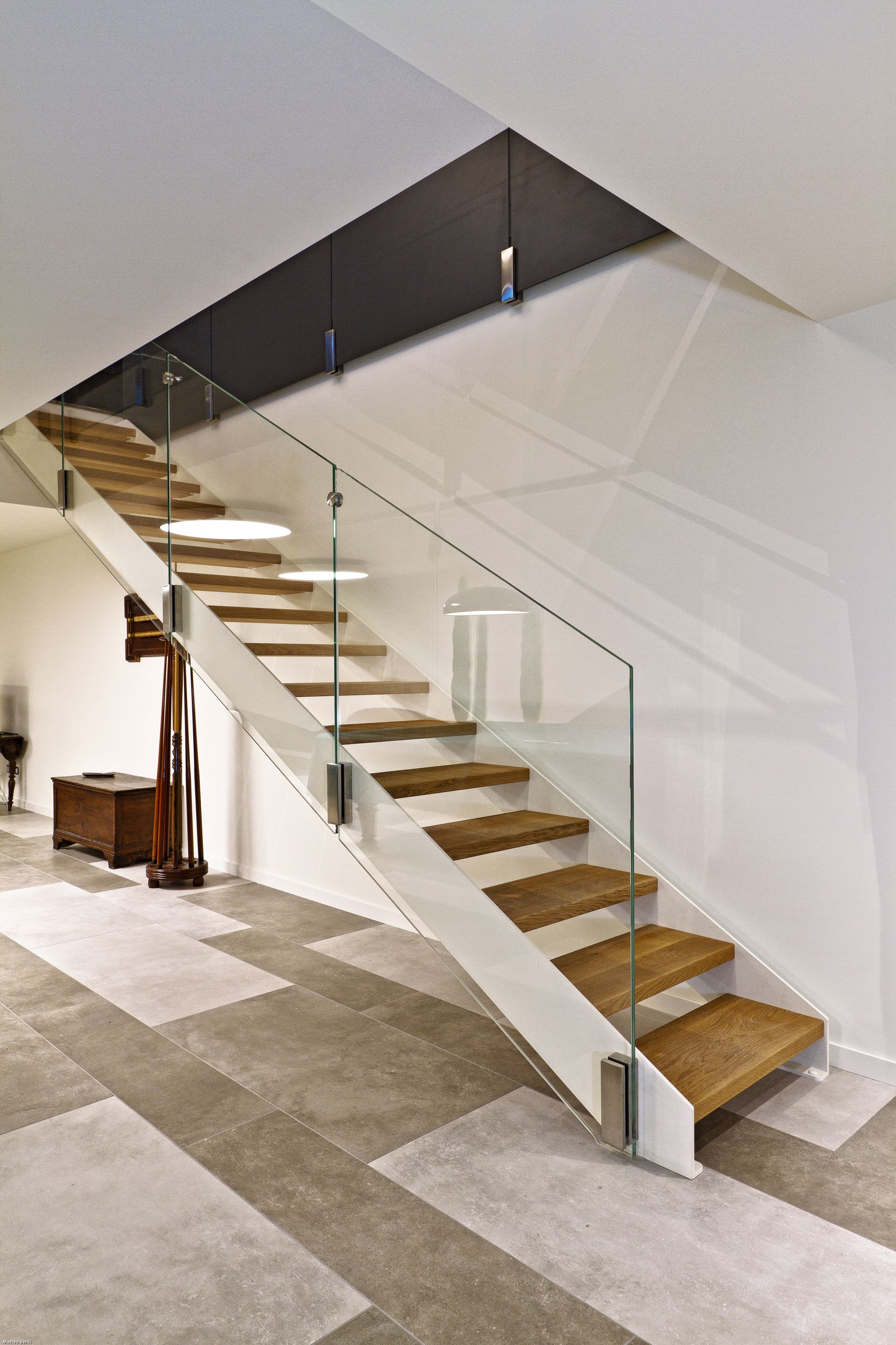 Scala interna moderna con gradini in legno, ringhiera in vetro e moquette grigia su sfondo bianco.