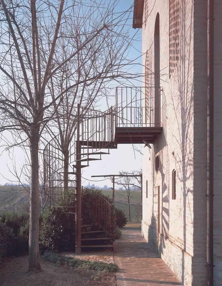 Scala esterna in mattoni con pianerottolo e ringhiera in metallo, accanto a un edificio in pietra e alberi spogli.