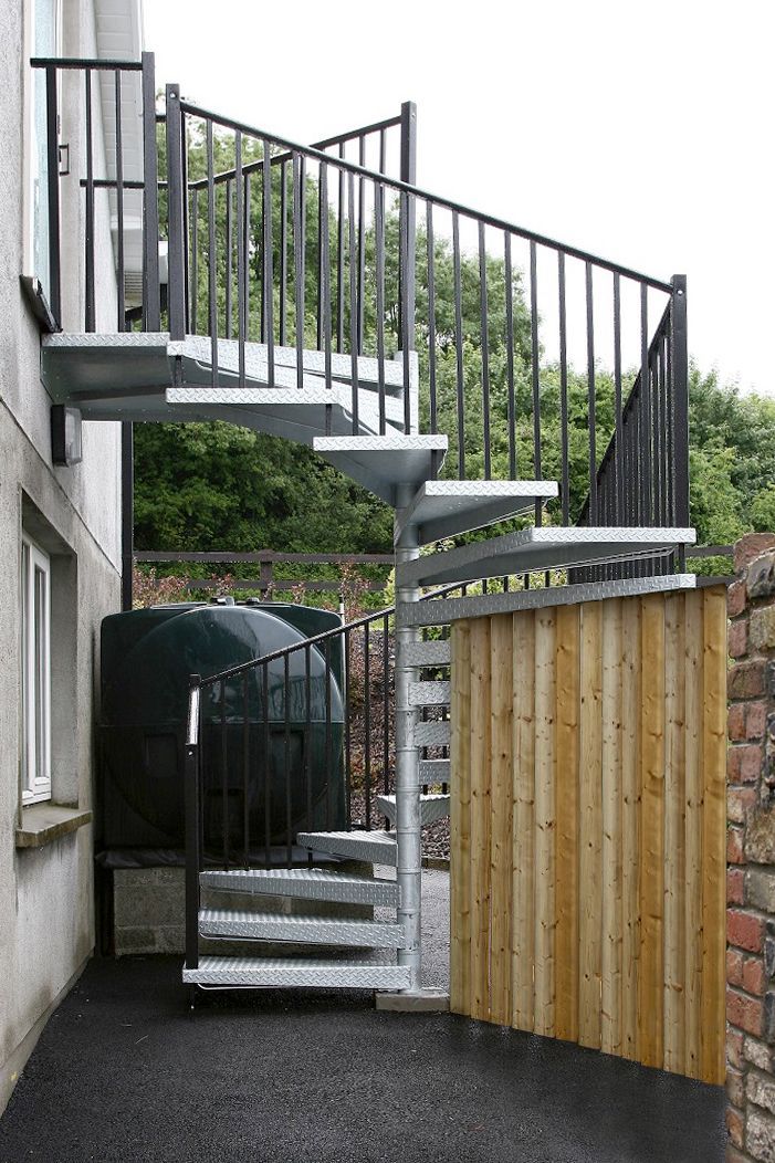 Scala a chiocciola esterna in metallo accanto a una recinzione in legno e al muro di una casa.