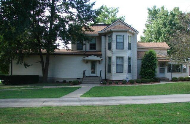 Two-story light-colored house with a rounded bay window, a small front porch, and a grassy lawn.
