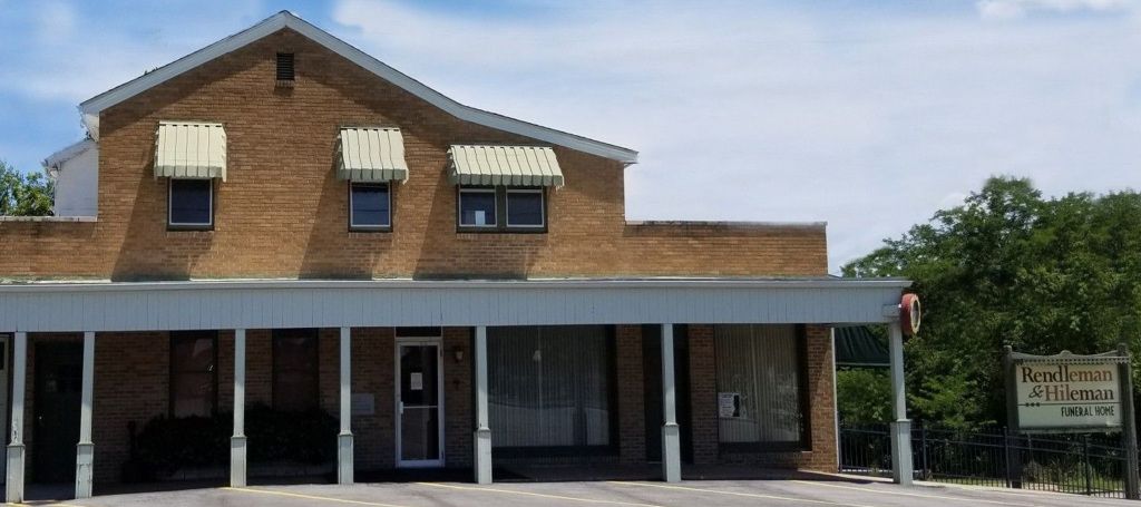 A brick building with awning over windows. A sign is to the right. Green trees surround.