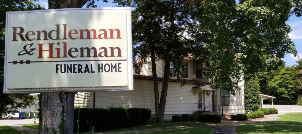 Sign for Rendleman & Hileman Funeral Home, brown text on a white sign, in front of the building, a sunny day.