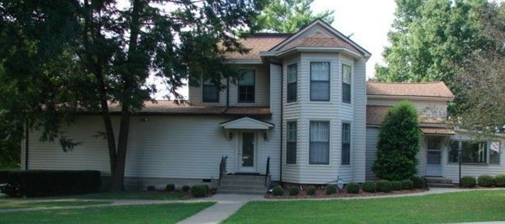 A two-story white house with a brown roof and a round turret. Trees and green grass surround the home.