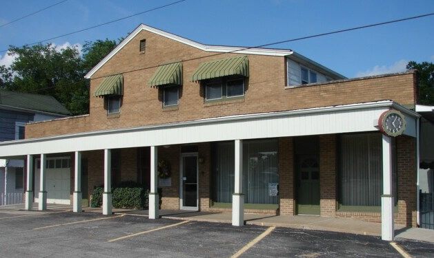 Two-story brick building with a covered porch and green awnings. Parking lot in the foreground.