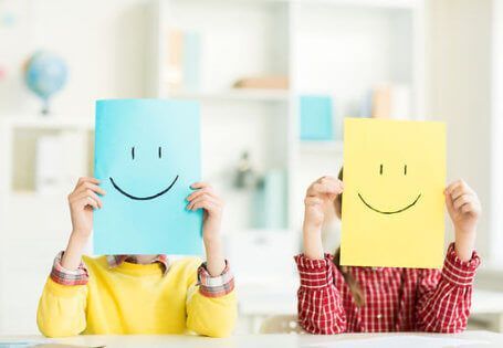 Two children holding blue and yellow smiley-face papers in a bright classroom