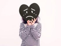 Person in a striped shirt holding a black heart-shaped sad face mask over their face.