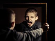 Child shouting at his reflection in a mirror, with a blurred figure in the foreground.