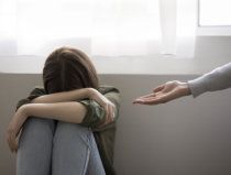 Person sitting curled up by a window while another hand reaches toward them
