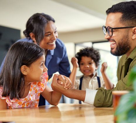 Two adults and two children arm wrestle and smile at a table indoors.
