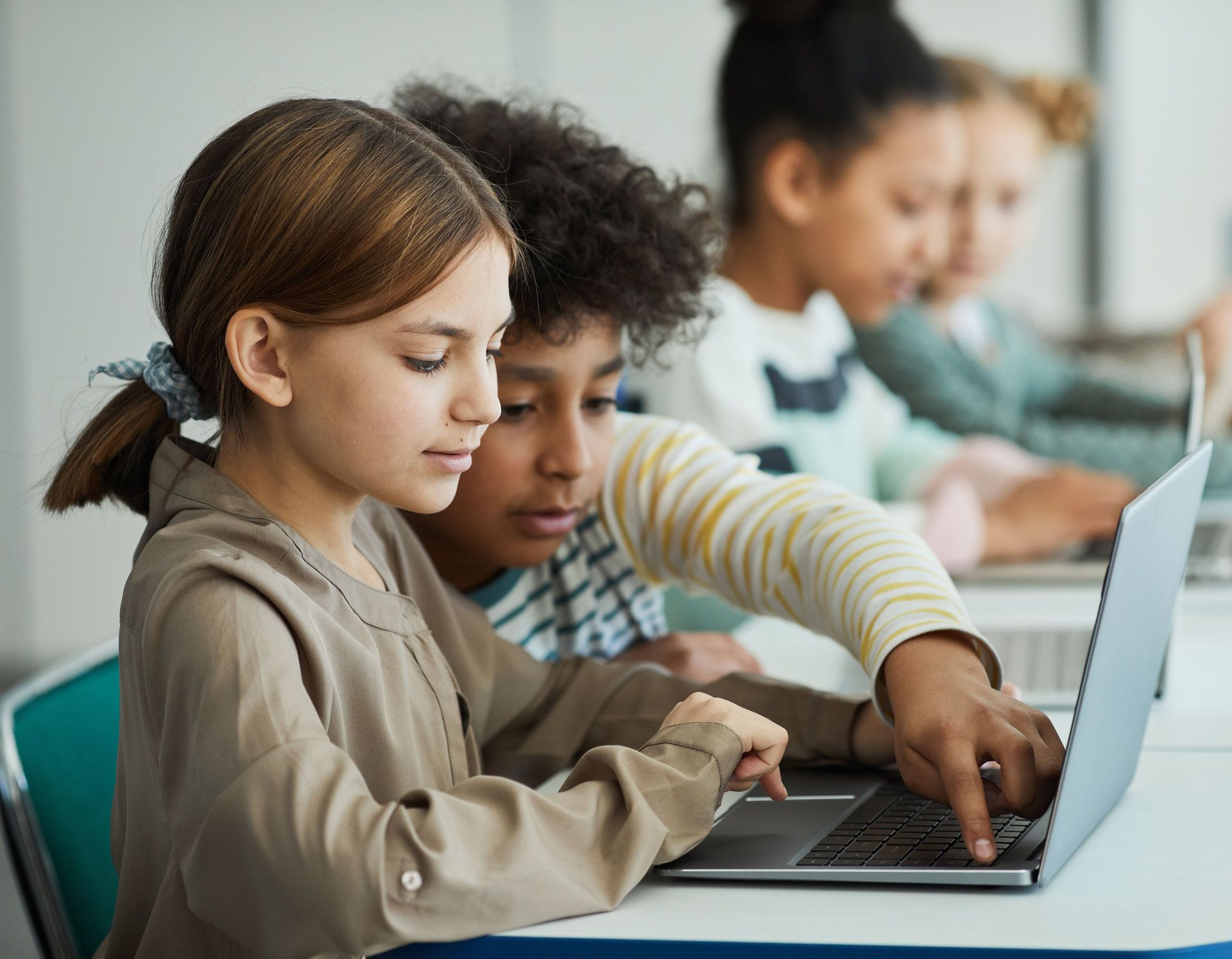 Children working on laptops at desks. One child points at the screen. Classroom setting.
