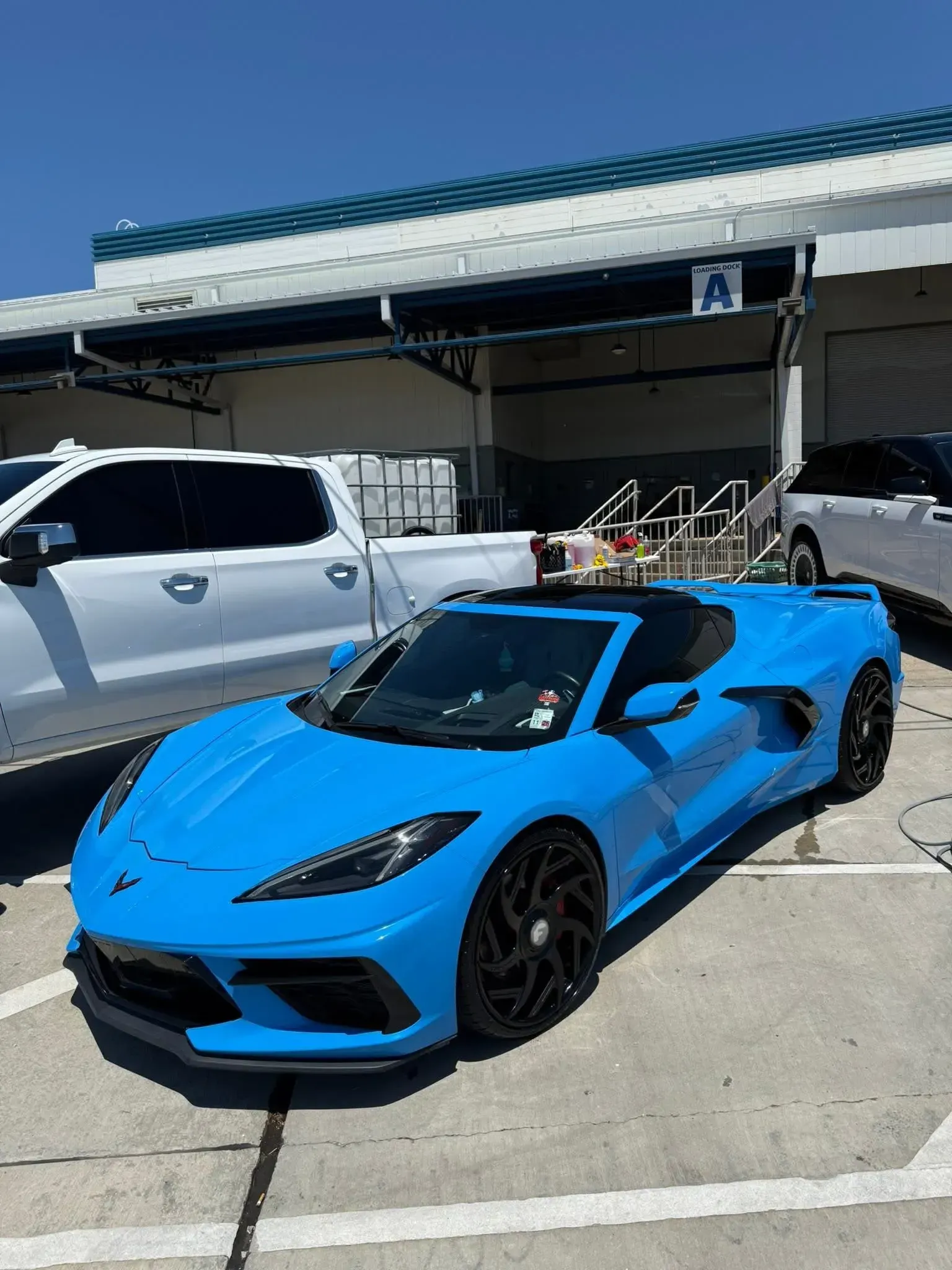 Bright blue Chevrolet Corvette convertible parked in front of a building with a white truck.