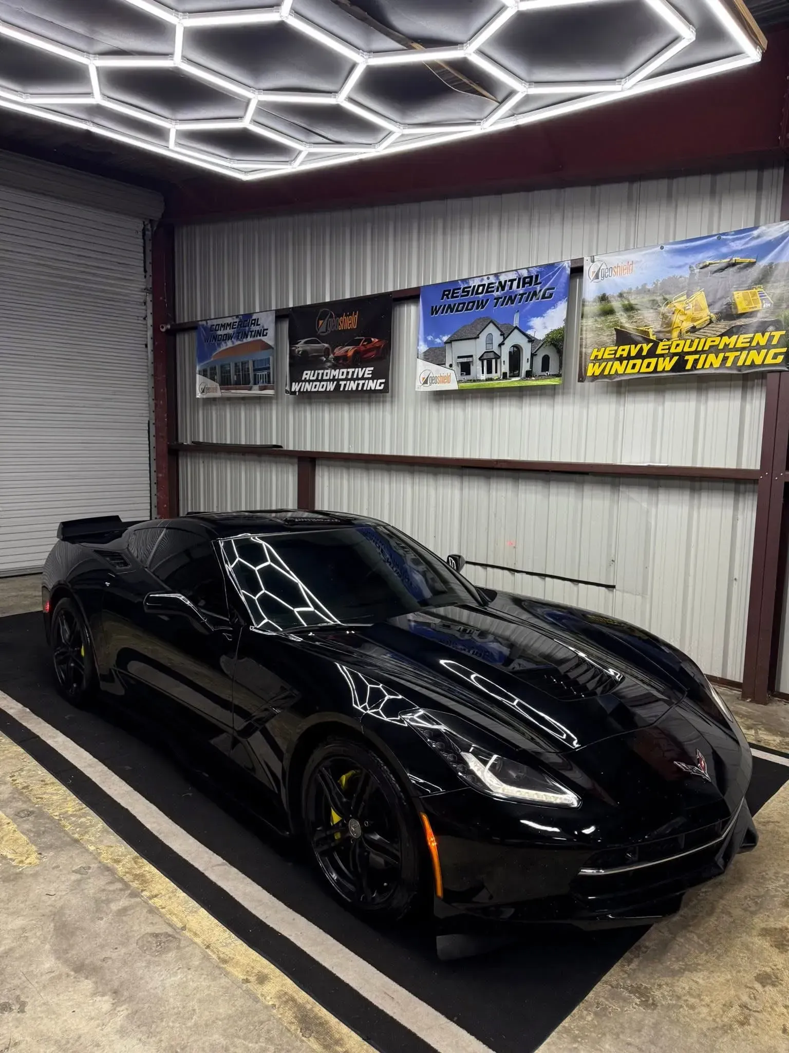 Black Corvette in a garage with hexagon-shaped lights. Garage has advertising posters hanging on the wall.