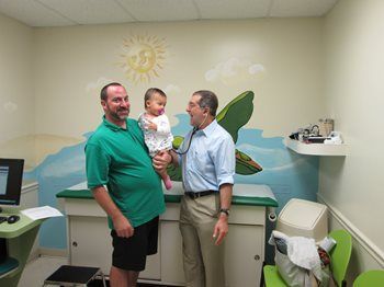 Father holding baby standing next to pediatrician with stethoscope.
