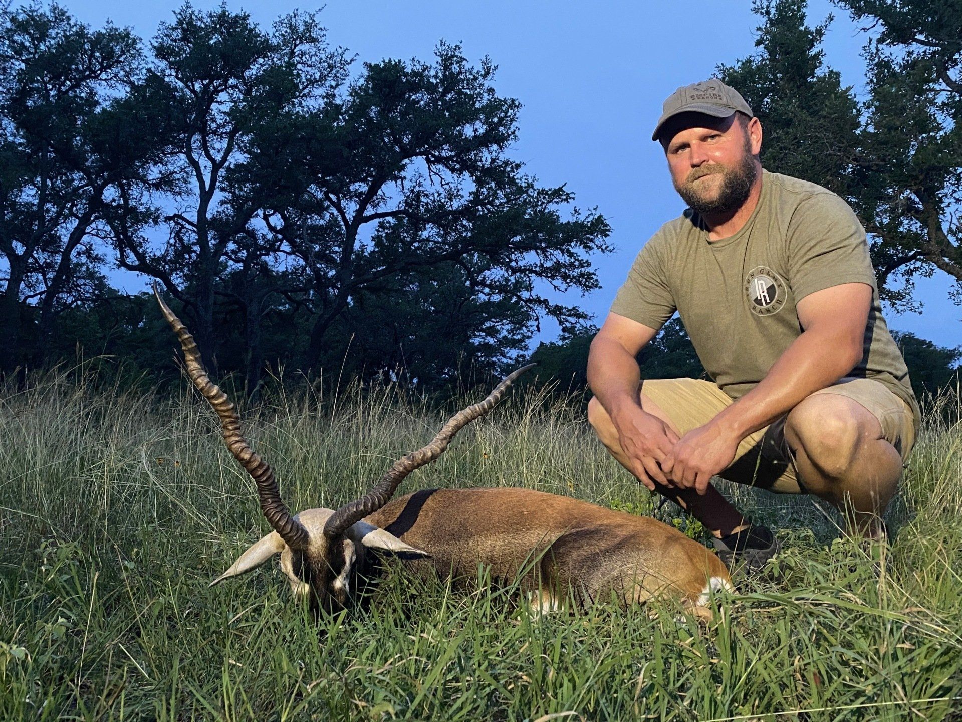 Man kneels next to a dead blackbuck antelope in grassy field. He wears a hat and khaki shorts, smiles.