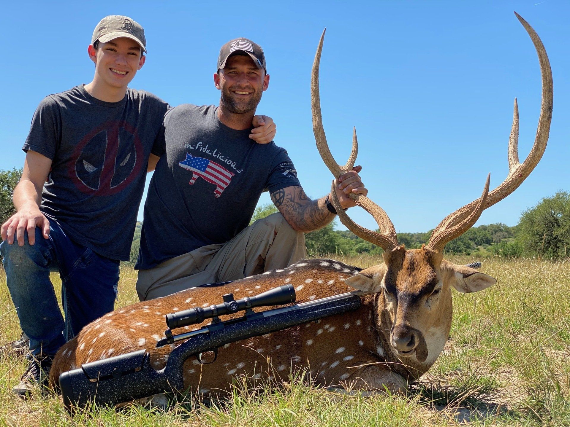 Man and teen pose with a spotted deer, rifle laid across it, outdoors. Sunny day.