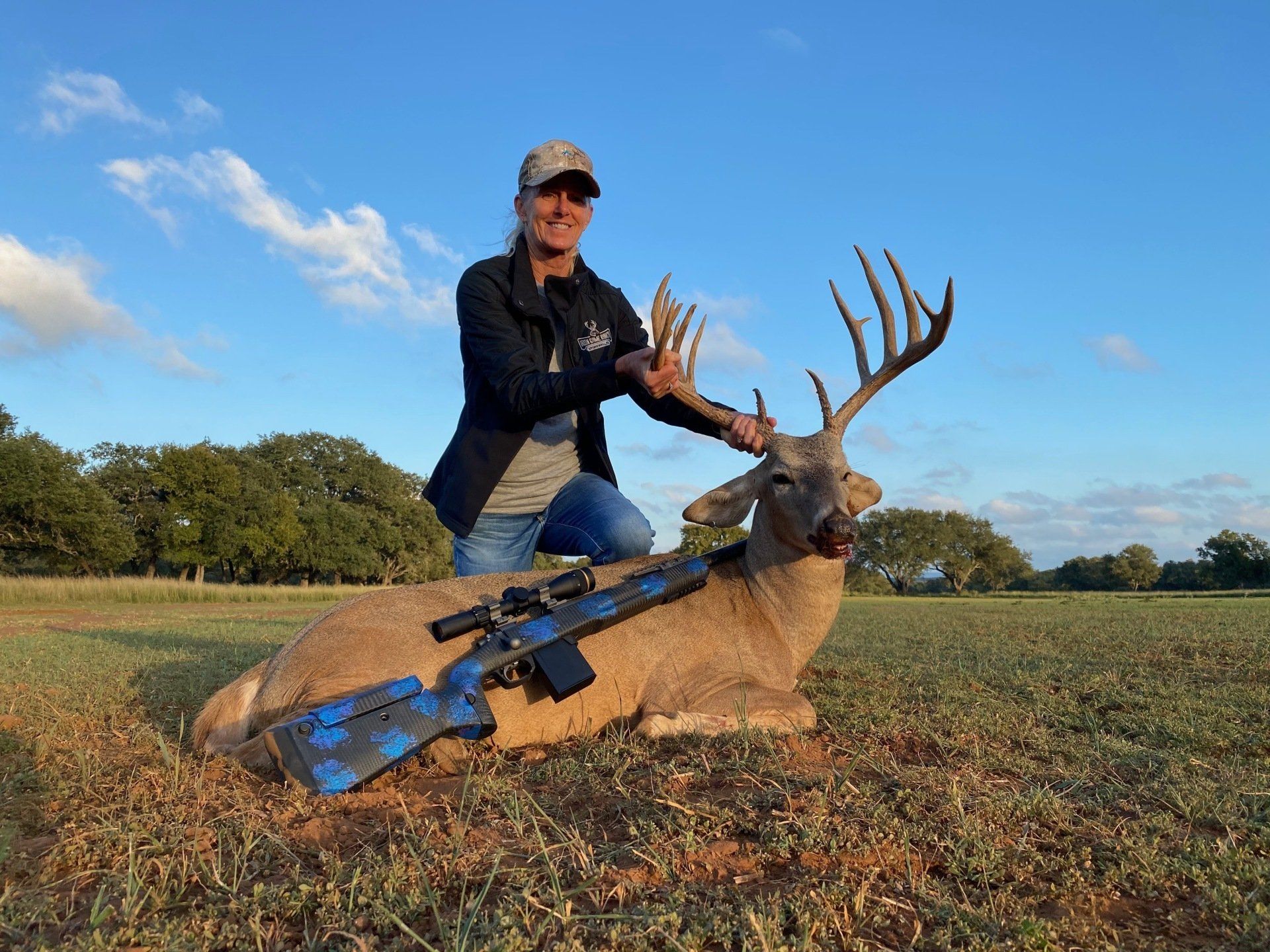 Woman kneels with a large buck she hunted in a field, holding its antlers, with a rifle lying beside.