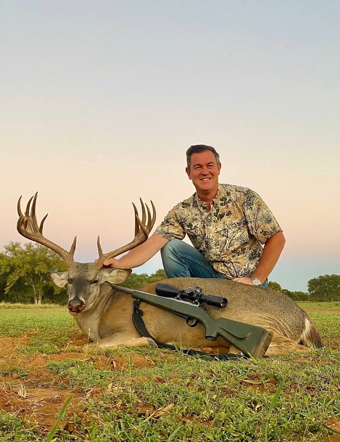 Man kneels next to a large buck he has hunted, holding its antlers. He smiles, rifle beside the deer in a field.