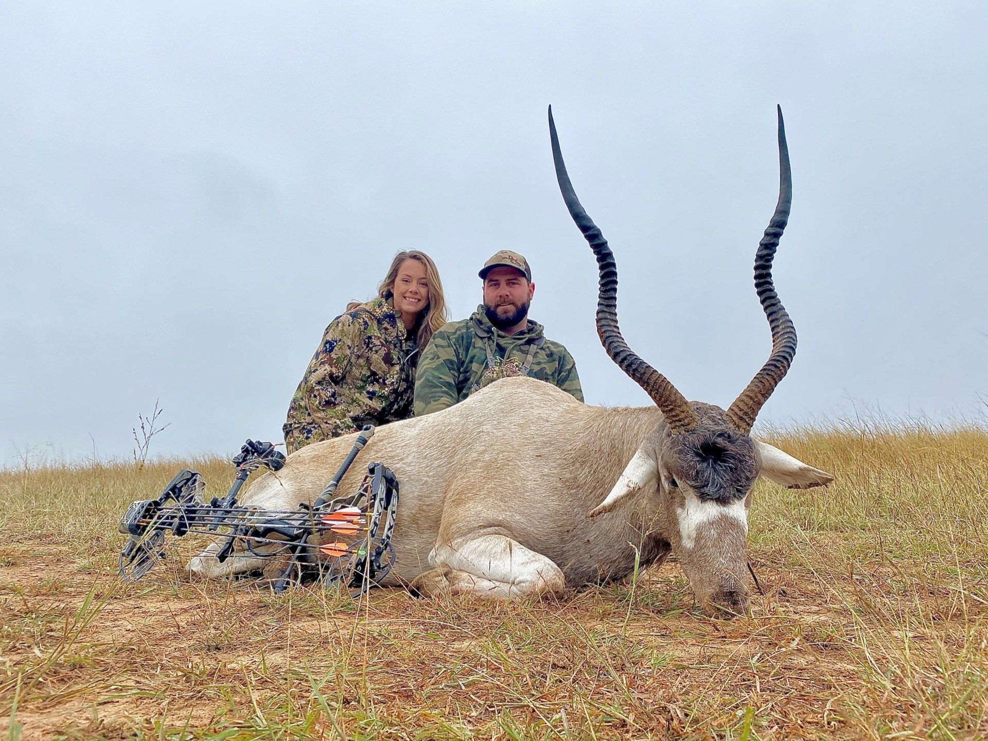 Two people pose with a hunted Addax. They are in camouflage, on a grassy field. Overcast sky.