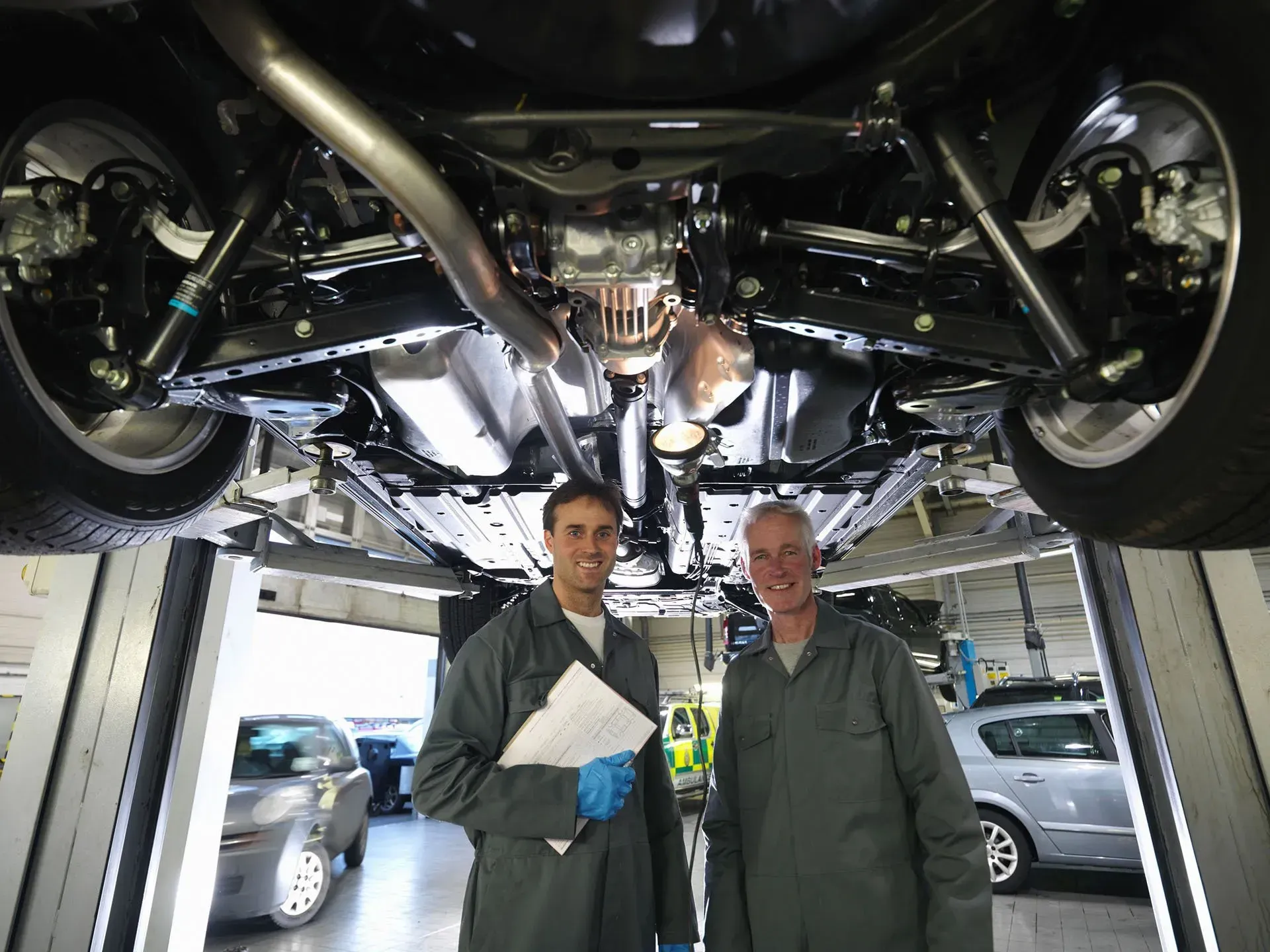 Two mechanics standing under a raised car in an auto repair shop, inspecting the underside.
