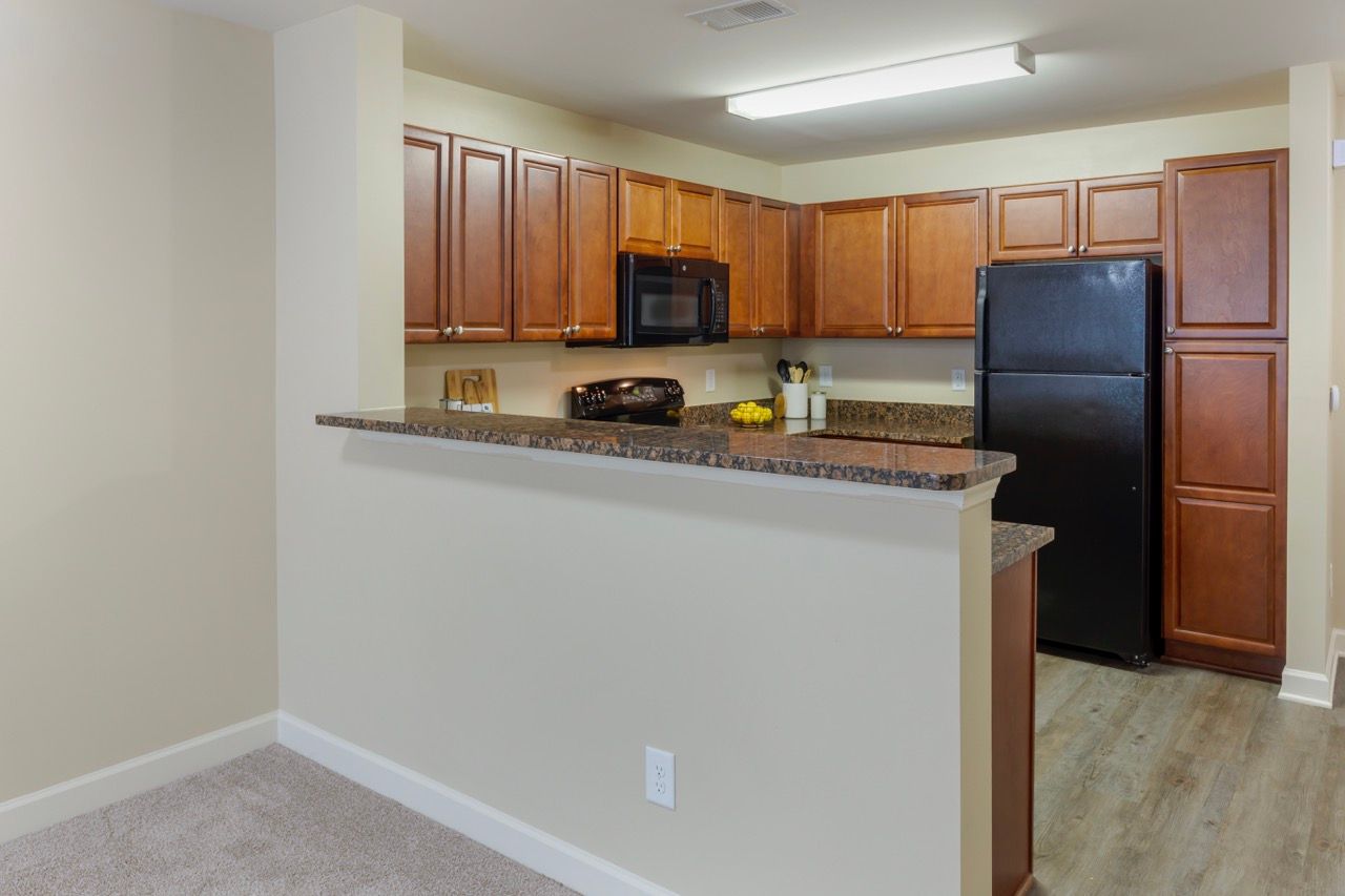 Open kitchen with brown cabinets, black appliances, and a bar counter in a beige apartment unit.