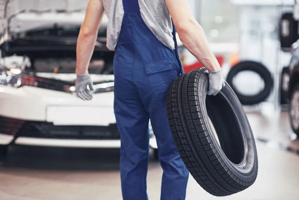 Mechanic in Blue Overalls Carrying a Tire in a Garage, Vehicle in the Background — Wheel Alignment Centre in Braitling, NT