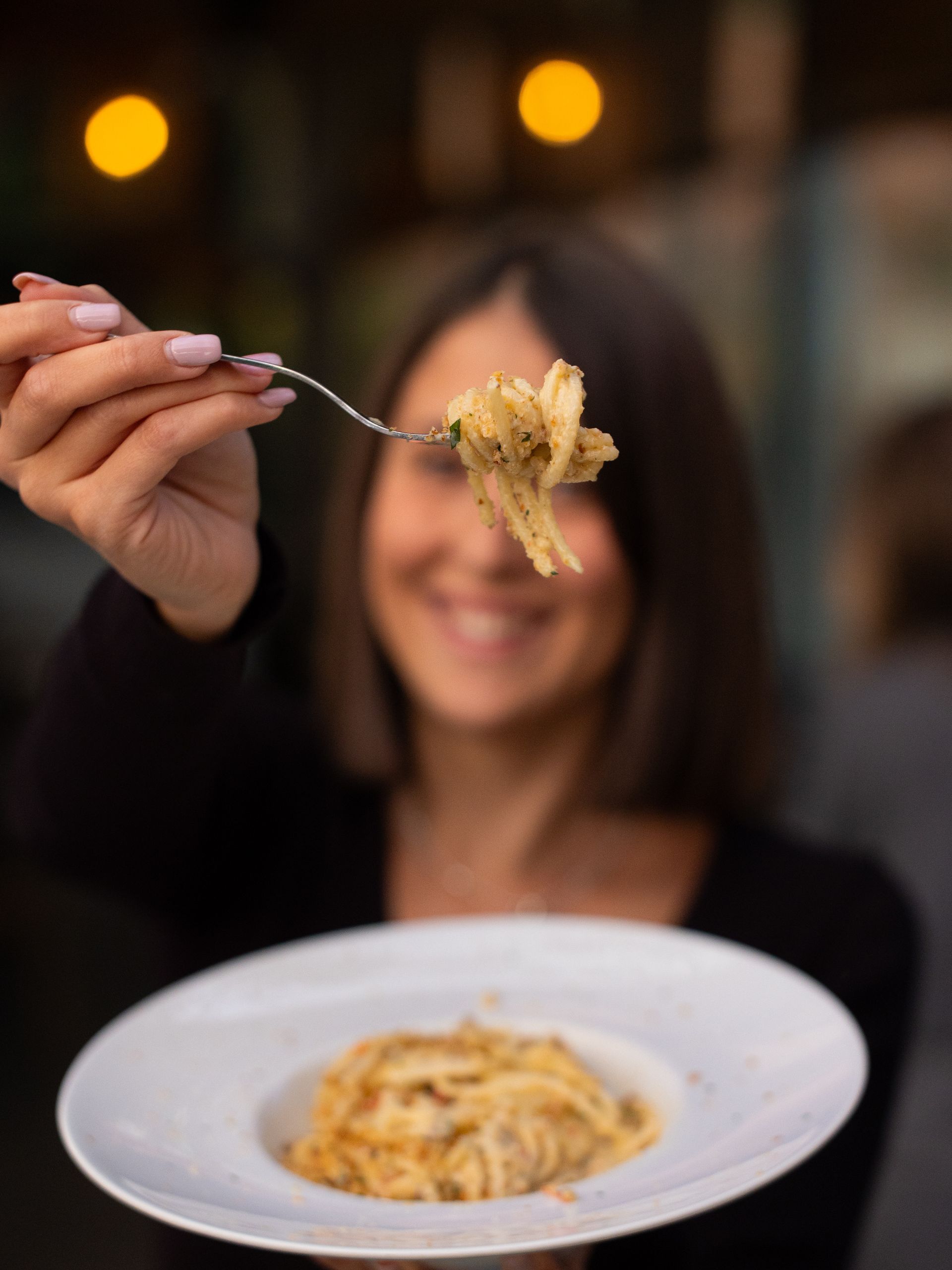 Donna sorridente che tiene in mano una forchetta con la pasta sopra un piatto di pasta. Sfondo sfocato.