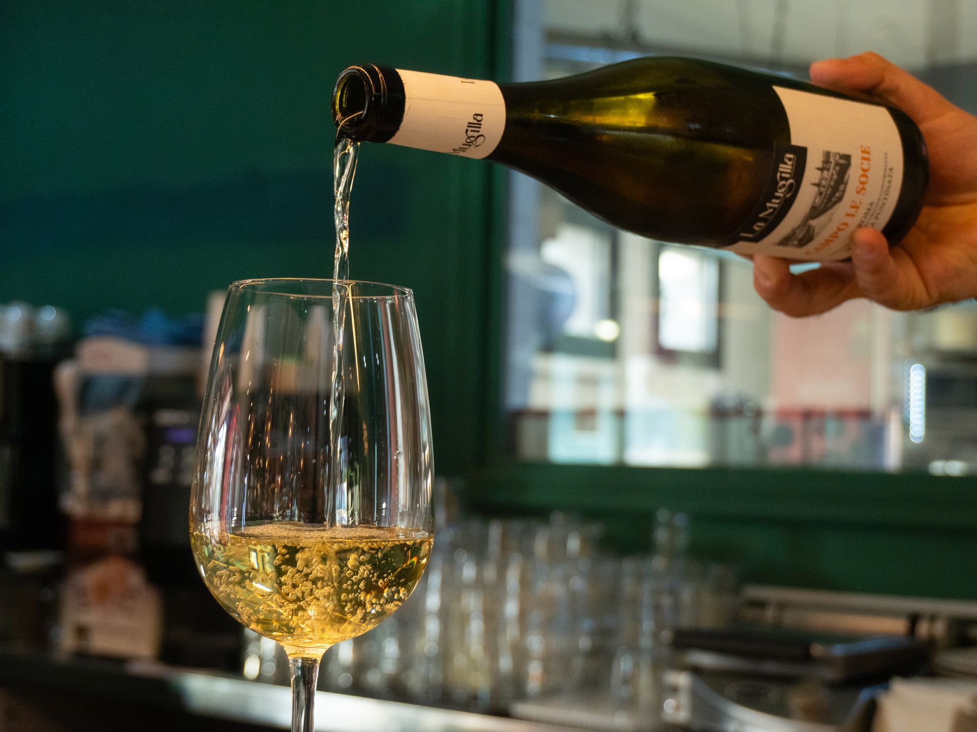 Una mano versa del vino bianco da una bottiglia in un bicchiere con stelo, in un ambiente da bar.