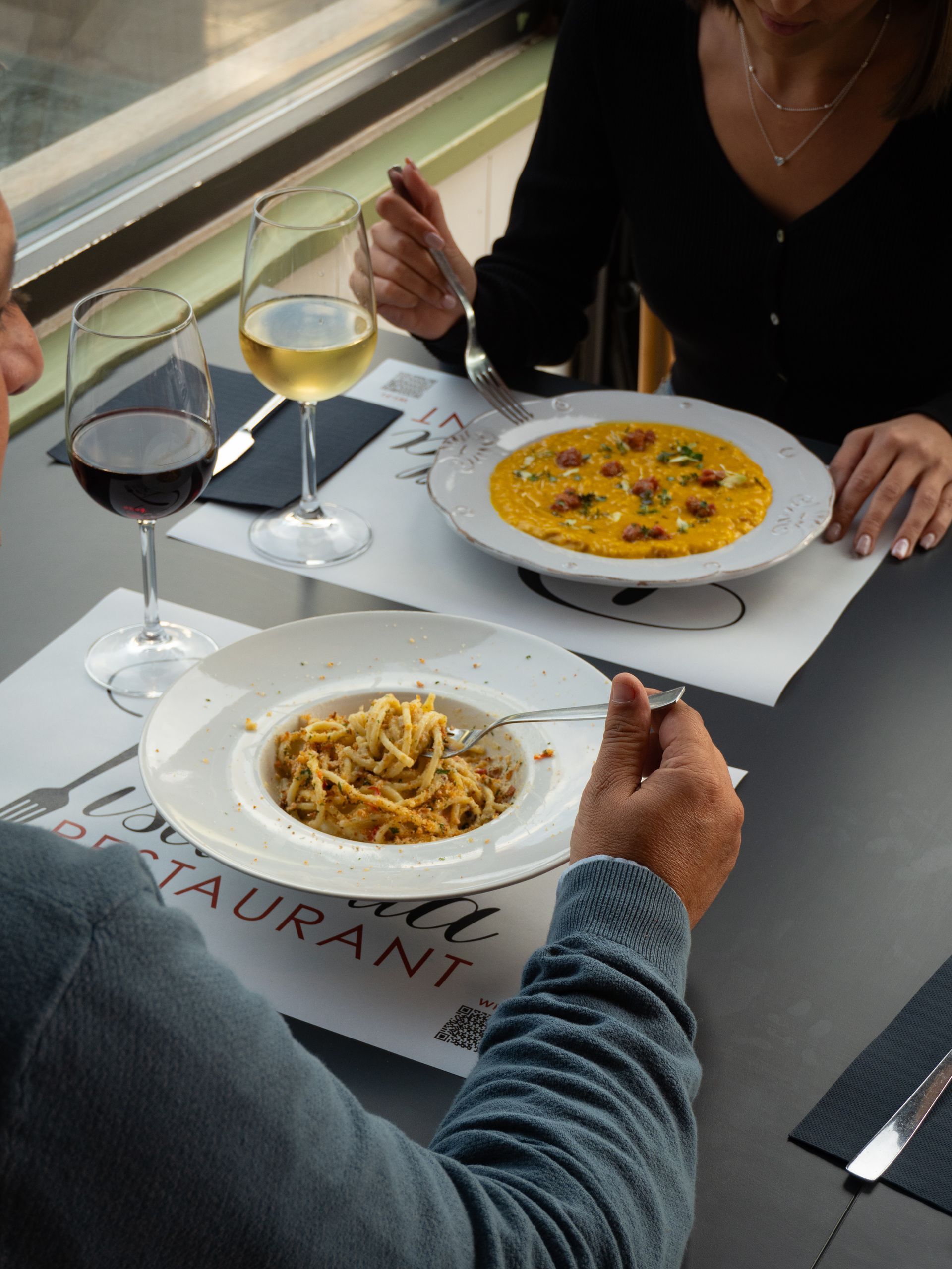Persone che cenano al ristorante: uno mangia pasta, l'altro riso. Bicchieri da vino e tovagliette sul tavolo.