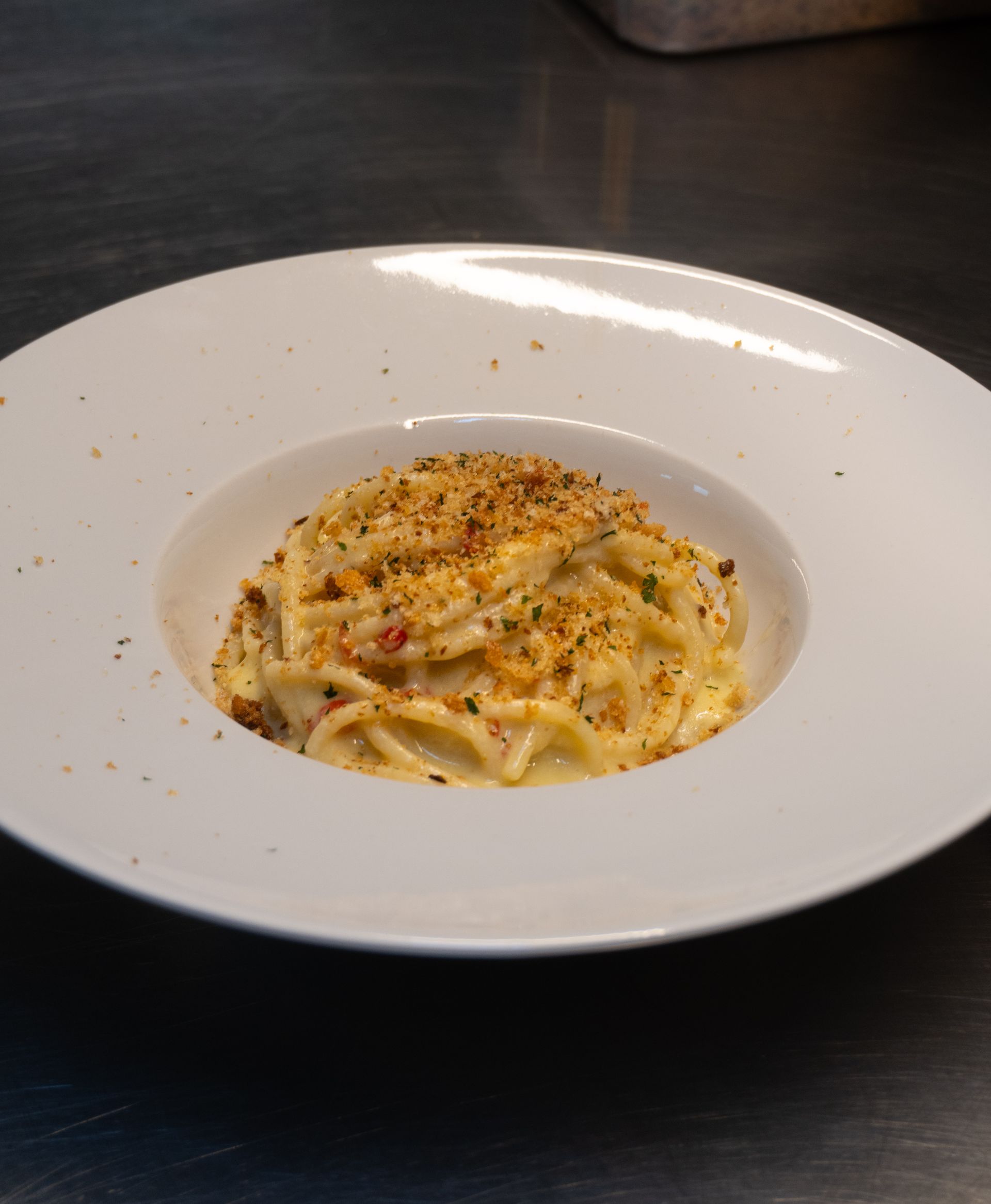 Piatto di pasta in una ciotola bianca, ricoperto di pangrattato, su una superficie scura.