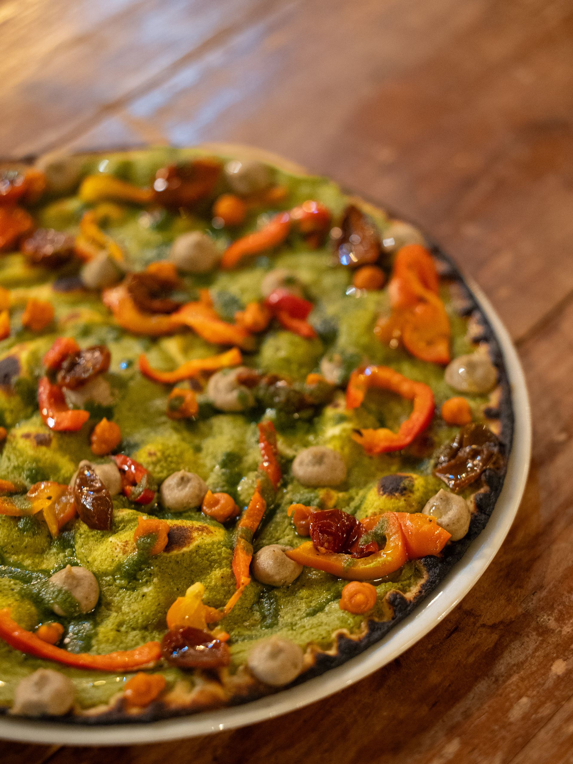 Pizza con salsa verde, peperoni arancioni e condimenti marroni su un piatto bianco sopra un tavolo di legno.