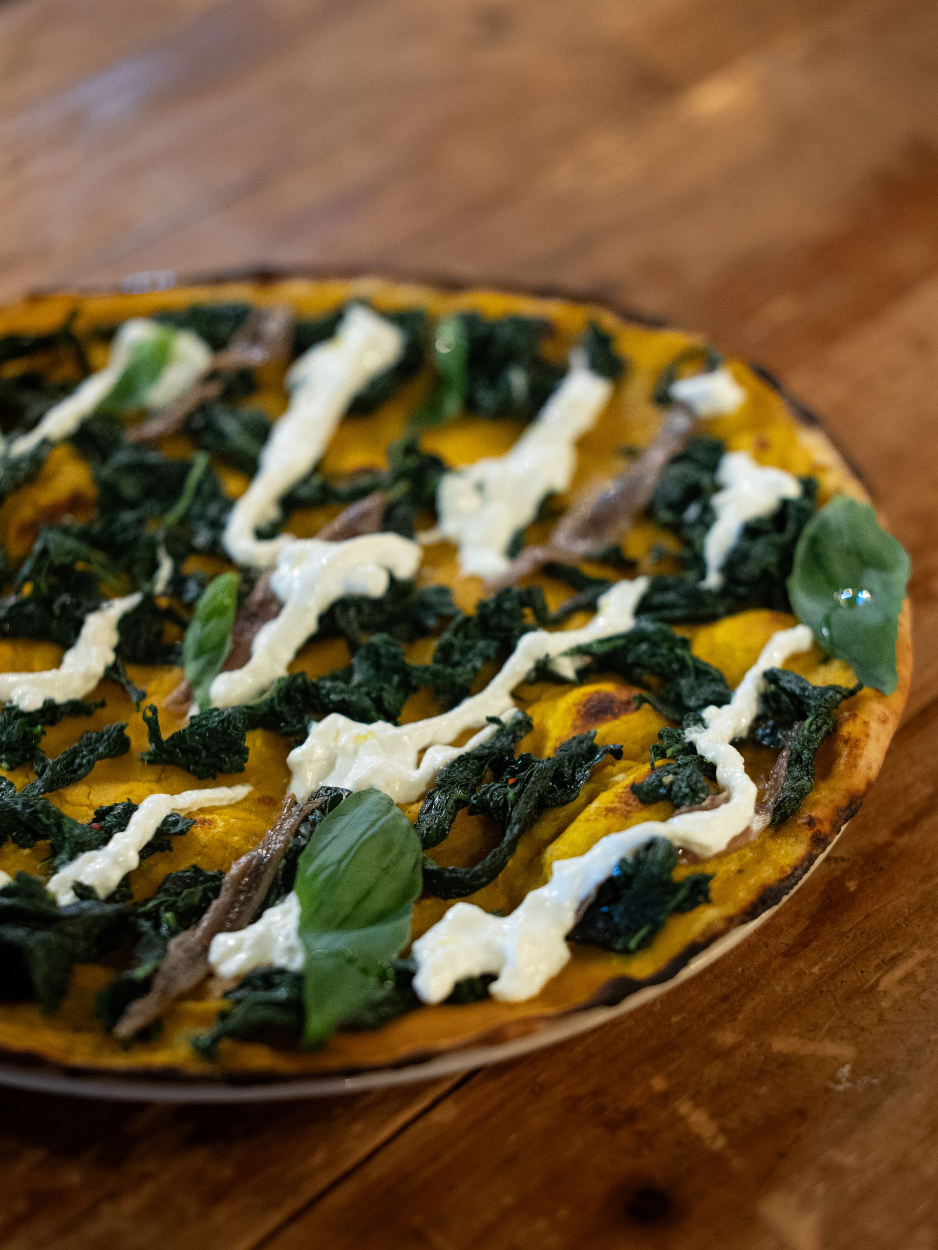 Pizza con base gialla, guarnizioni di foglie verdi, glassa bianca e acciughe su un tavolo di legno.