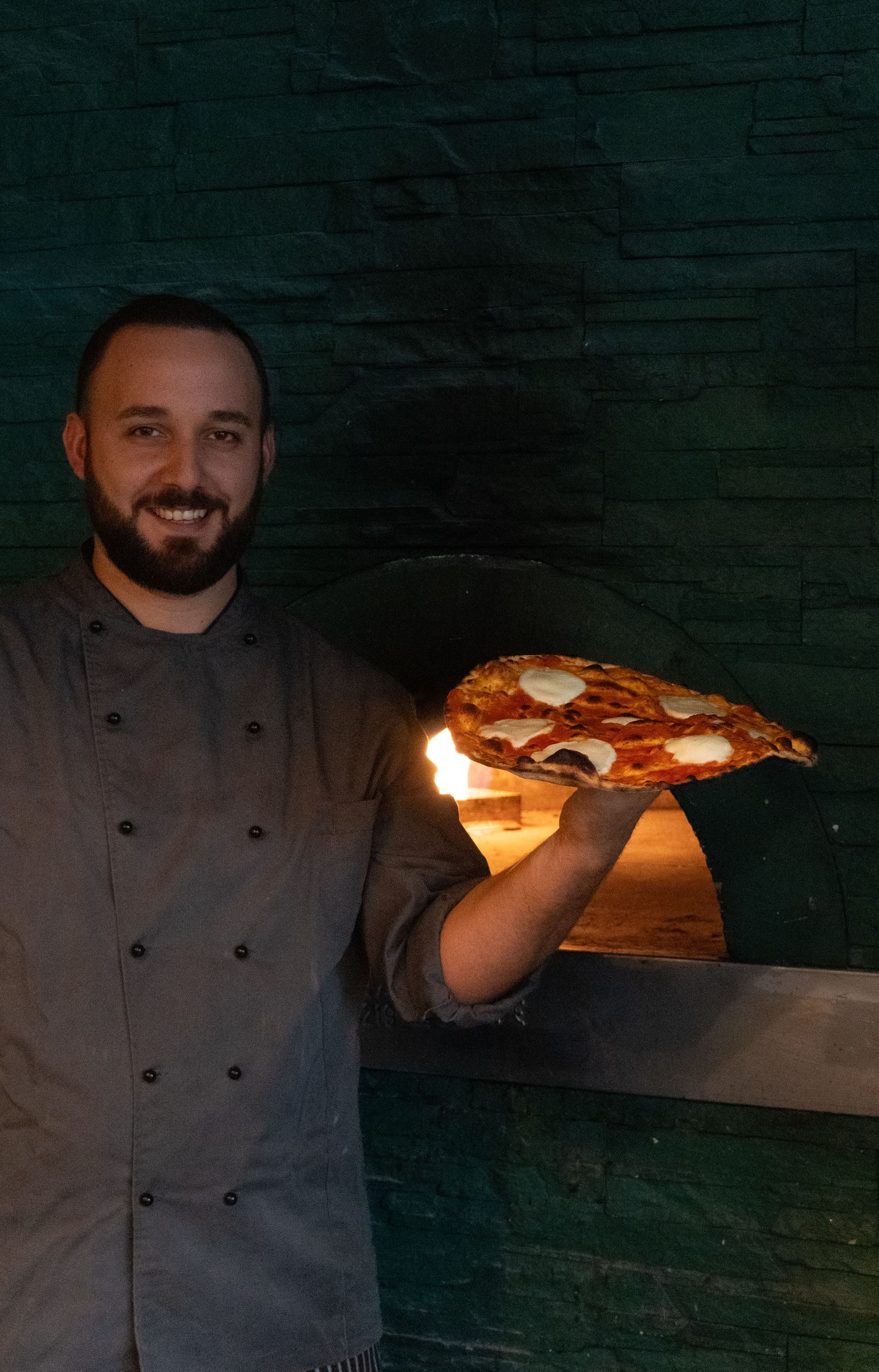 Cuoco che tiene in mano una pizza vicino al forno. Sorride in uniforme grigia con fiamme sullo sfondo.