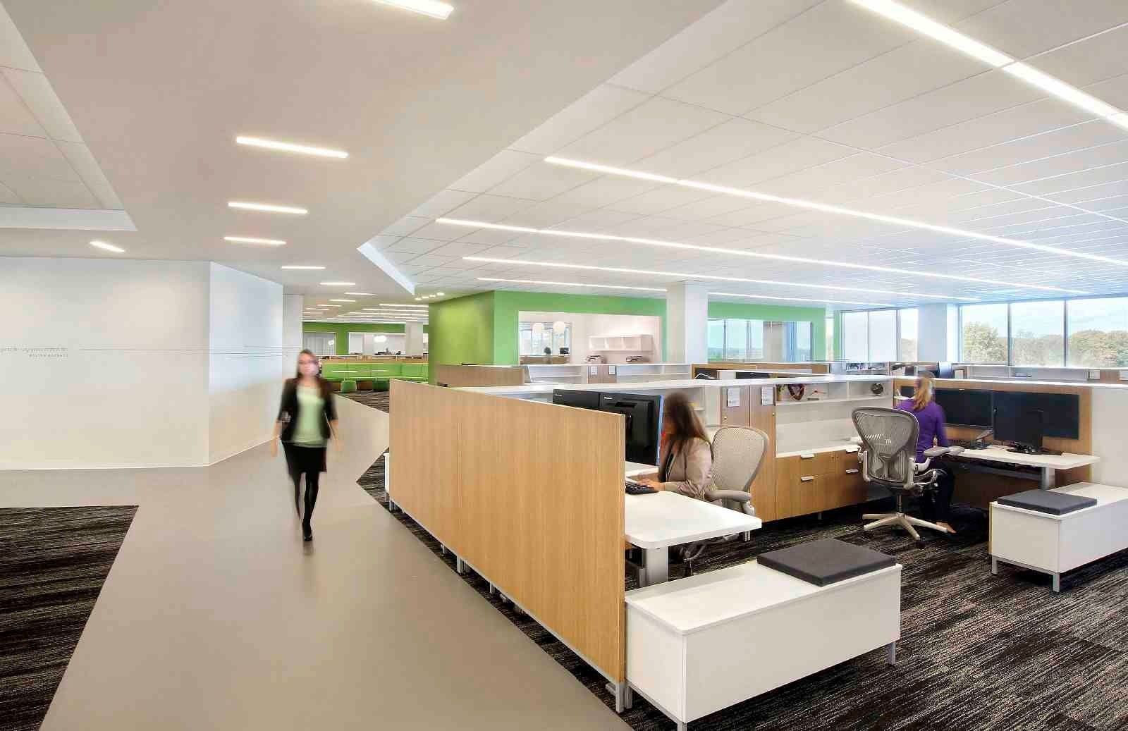 Moderno ufficio open space con dipendenti alle postazioni di lavoro e una persona che cammina in primo piano. Bianco, legno e dettagli verdi.