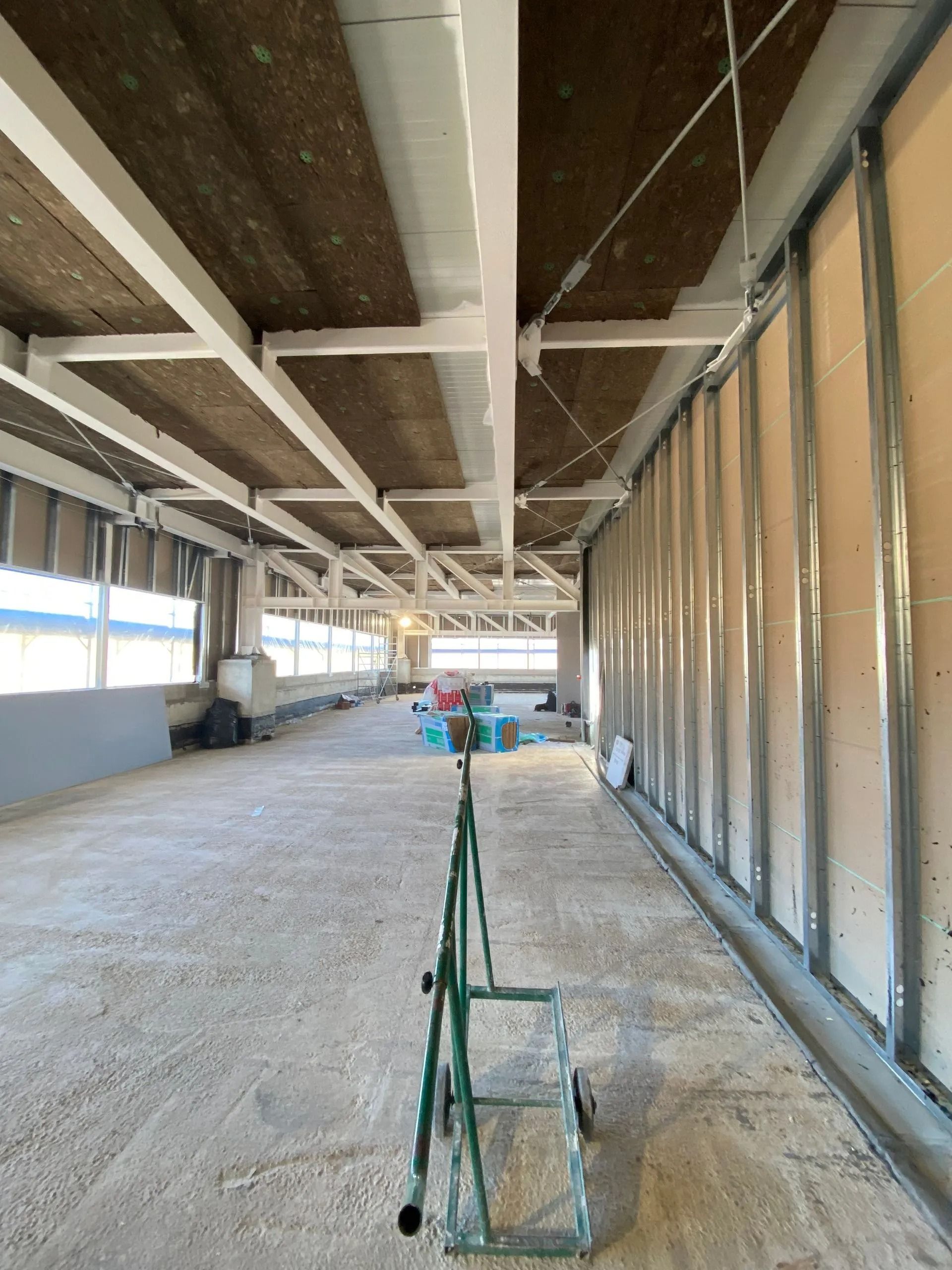 Cantiere interno con struttura metallica, isolamento e pareti grezze. Carrello verde in primo piano.