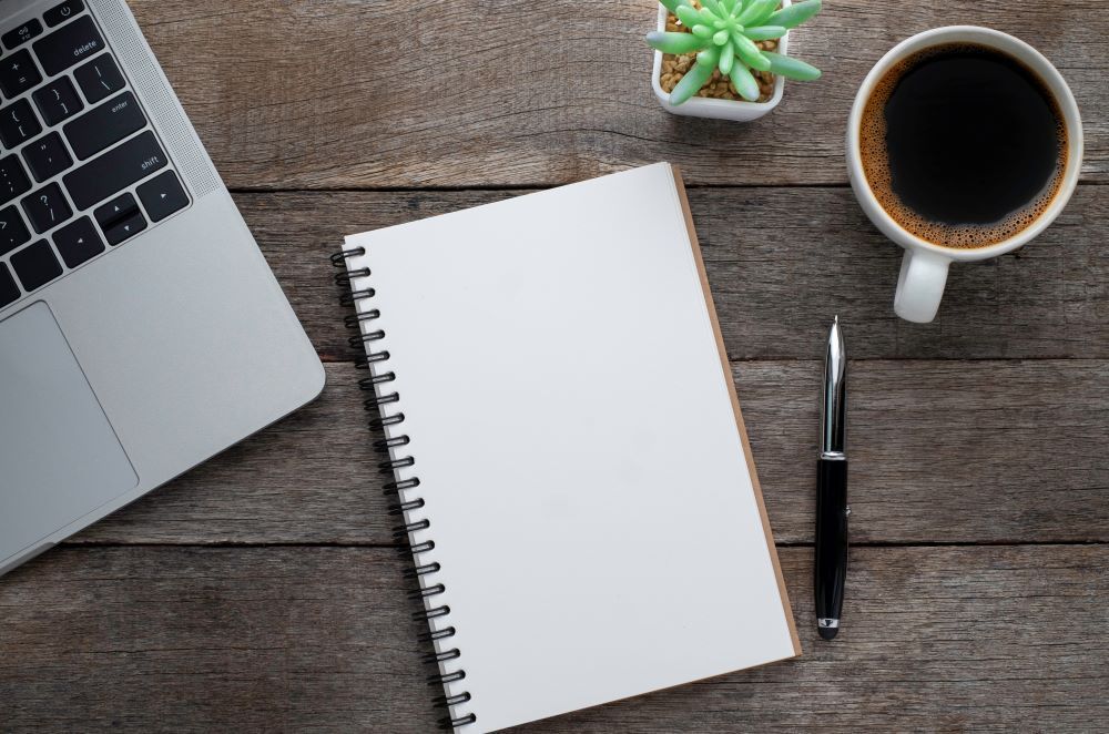 A notebook , pen , cup of coffee and laptop on a wooden table.