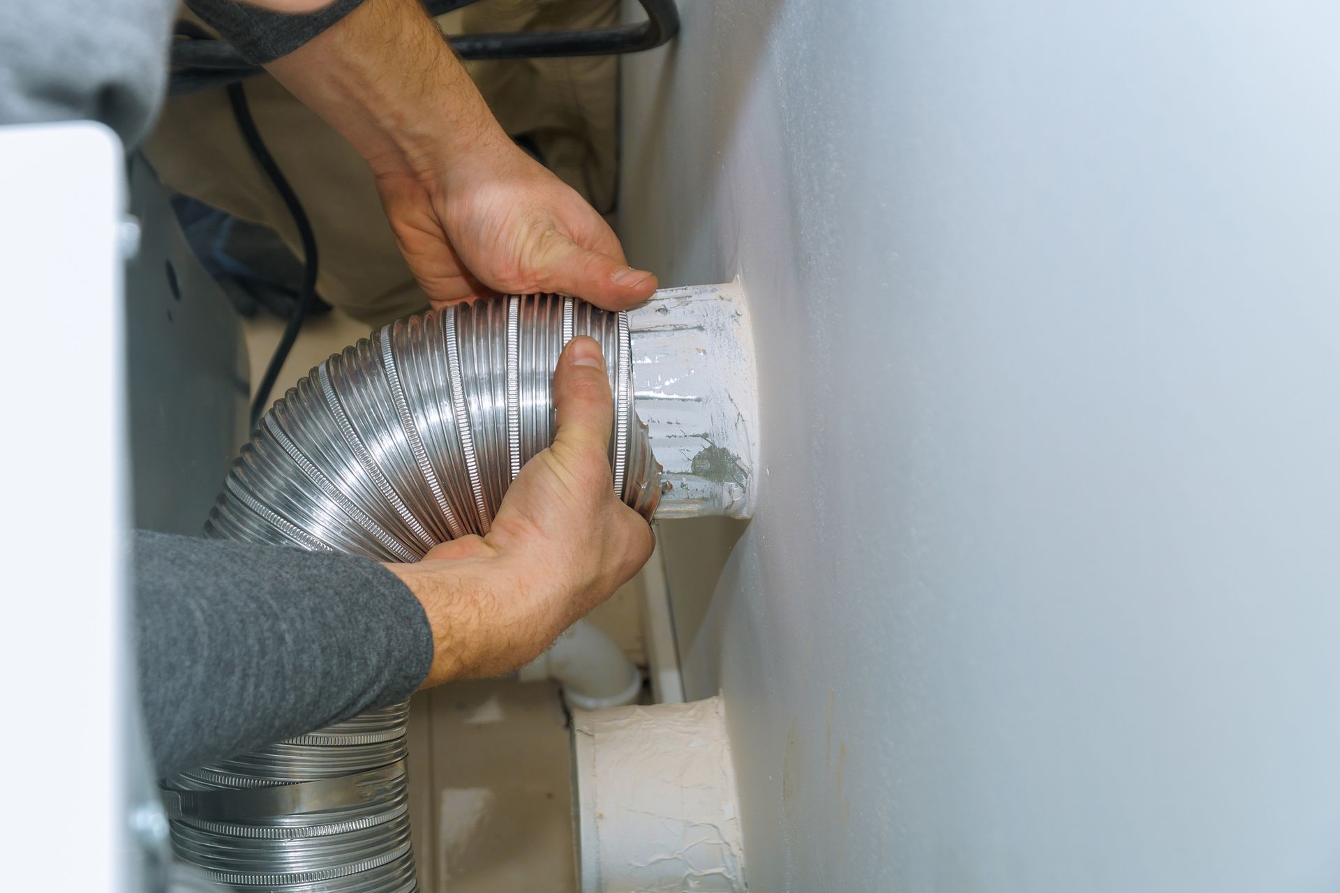 Hands connecting a silver dryer vent hose to a wall vent opening in a white room.