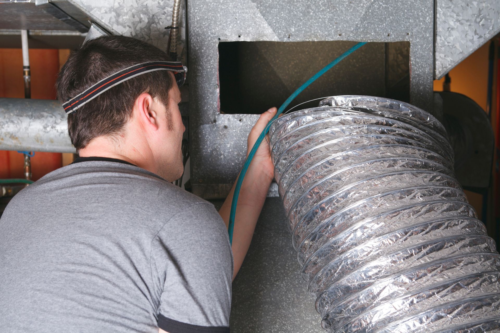 Man in grey shirt cleaning HVAC ductwork with a flexible brush, wearing a headlamp.