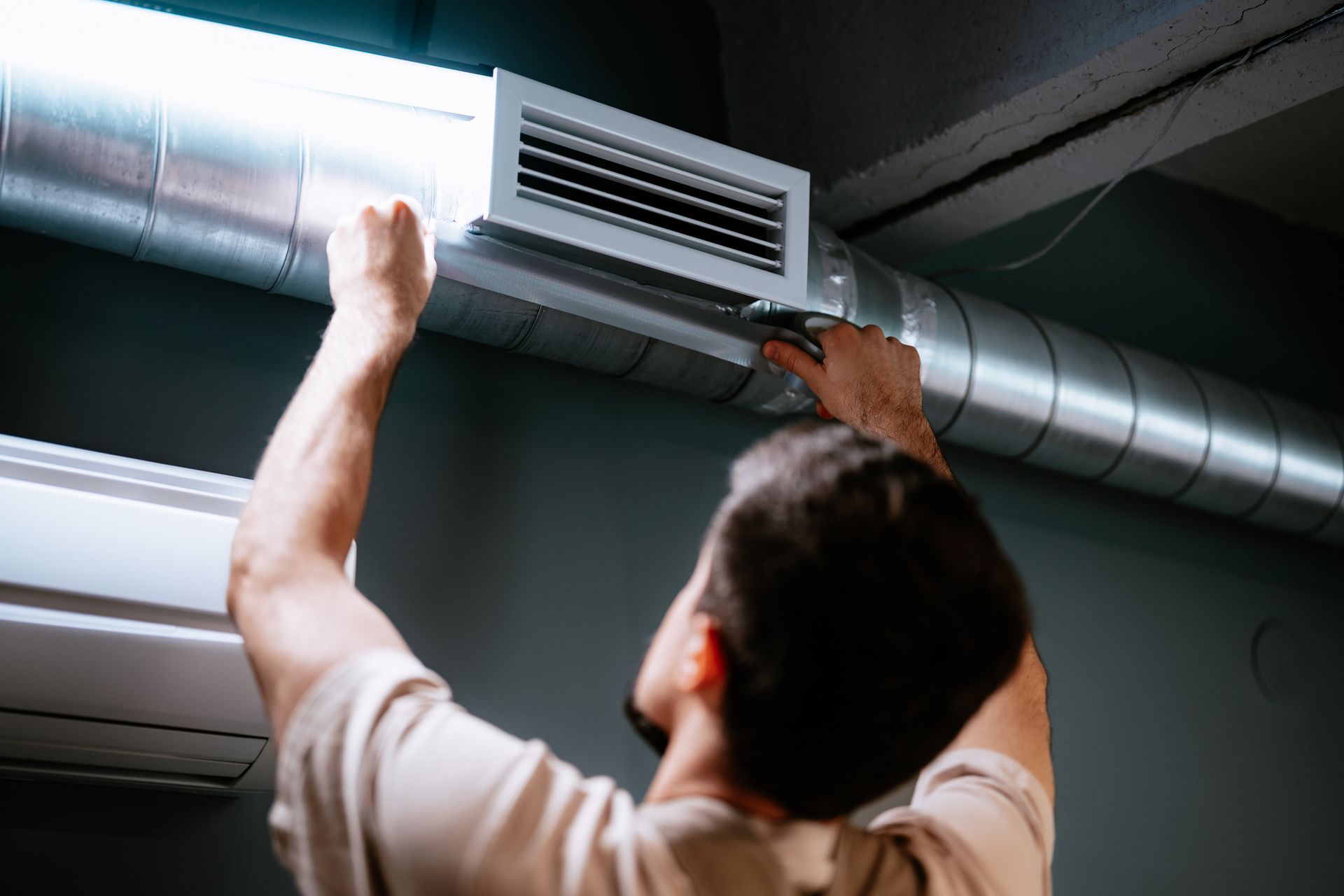 Person adjusting air vent on a duct. Gray wall, silver duct, white AC unit.