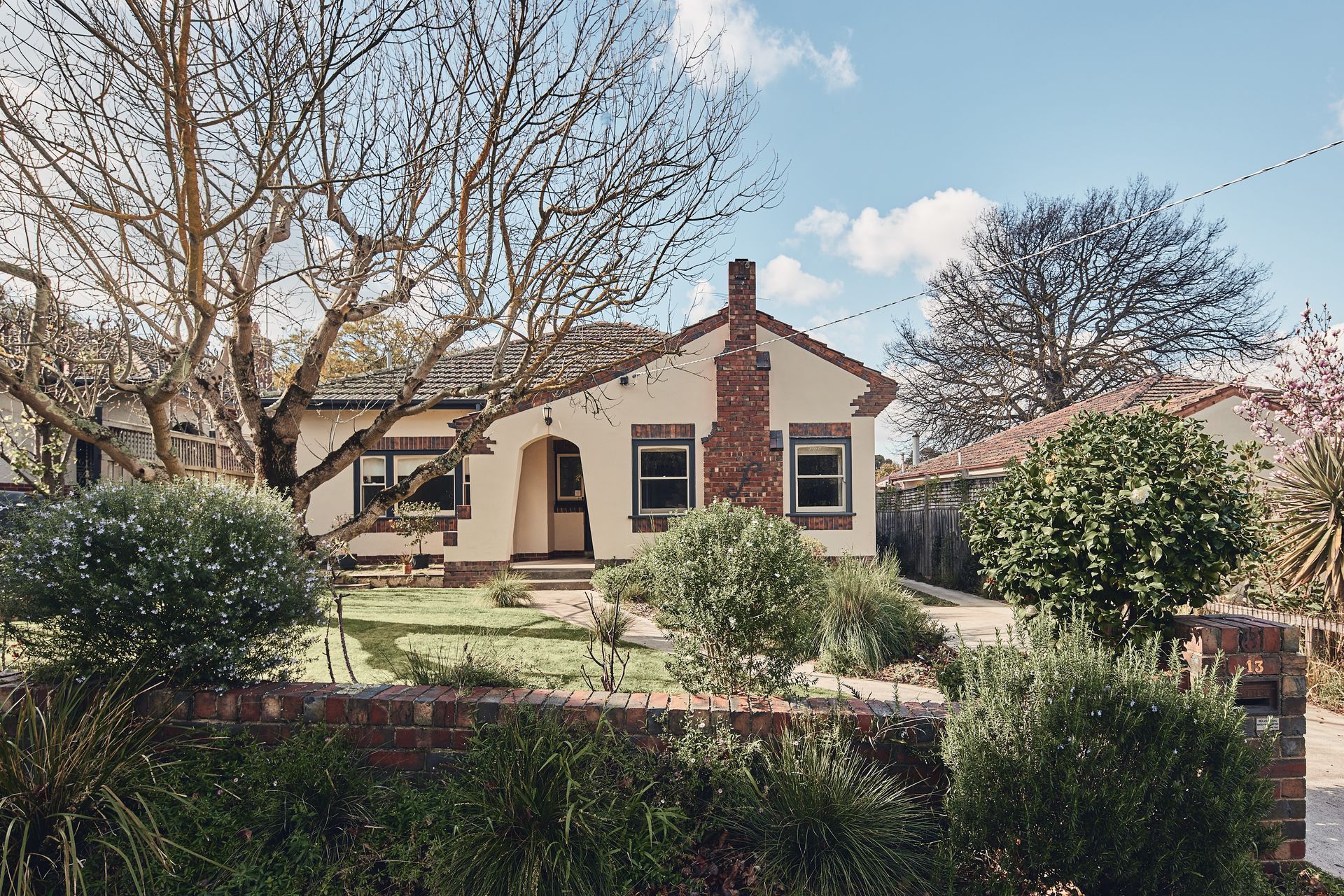 A beige house with a brick chimney and a small garden in front. Two trees and bushes are in the yard, under a blue sky.