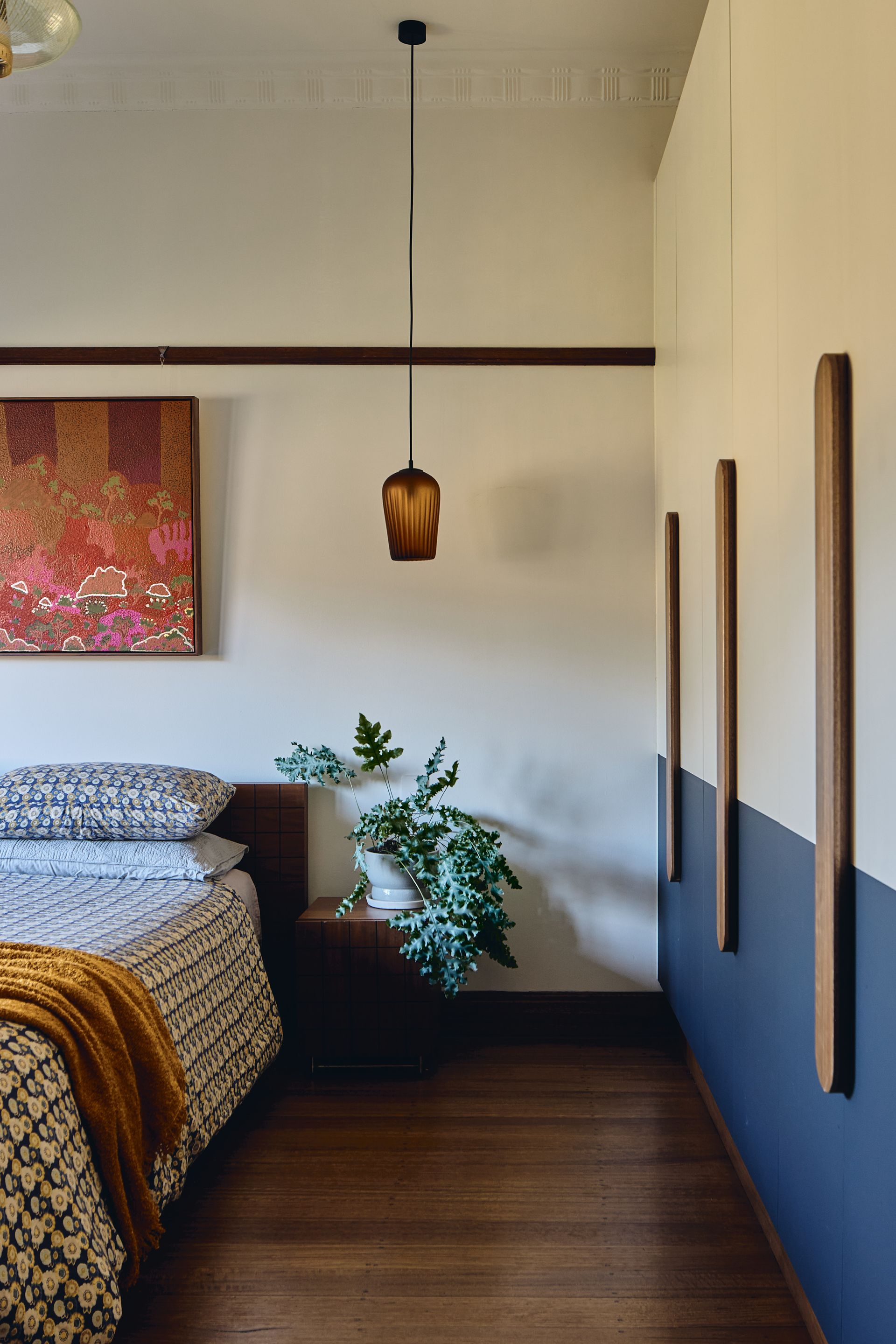 A bedroom with a bed , nightstand , plant and painting on the wall.