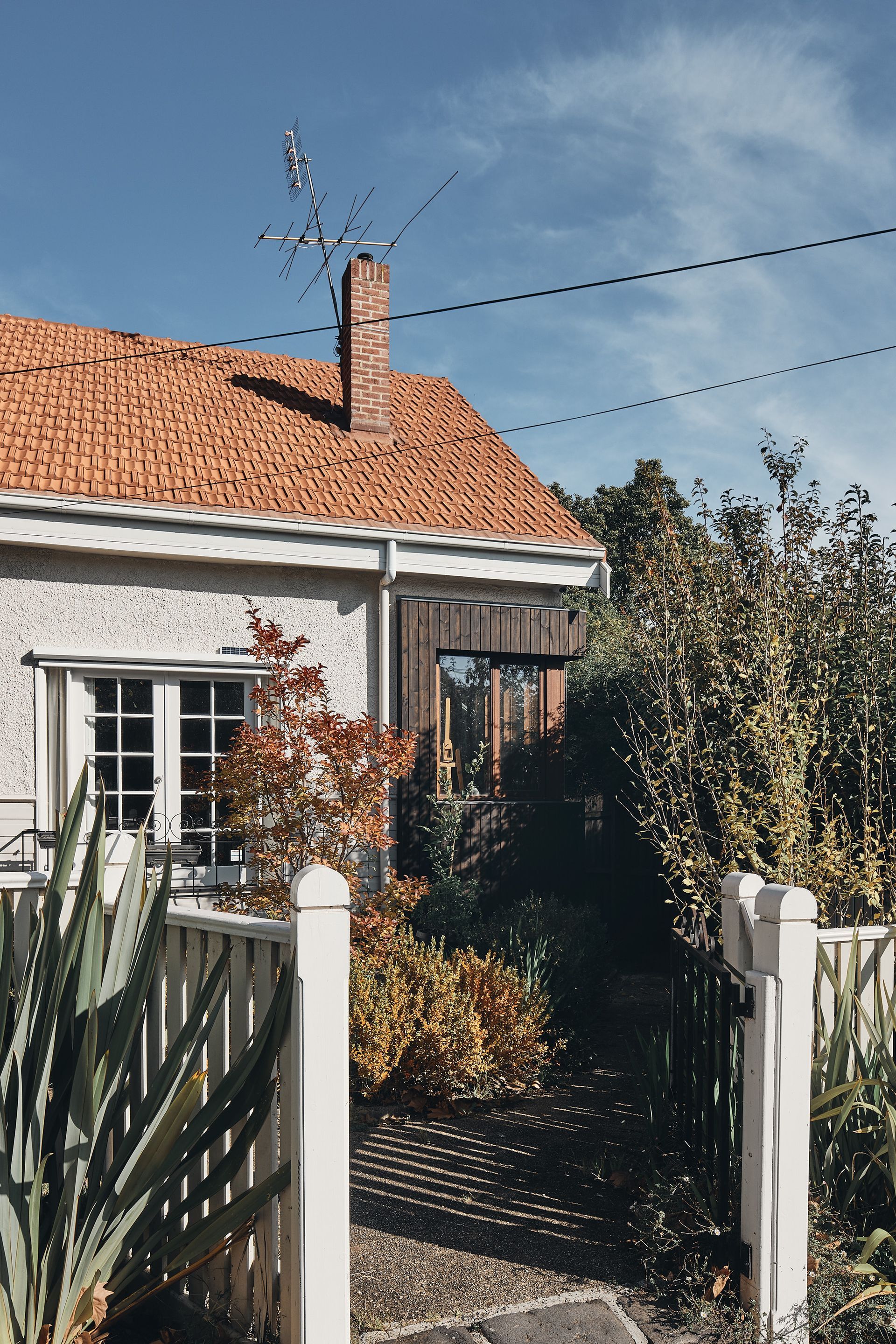 A house with a brick roof and a chimney is behind a white fence.
