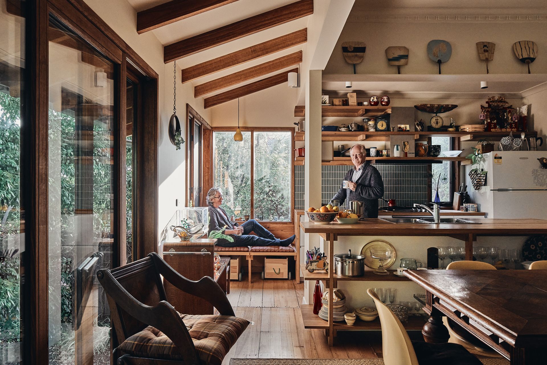 A man is standing in a kitchen while a woman sits on a bench in a living room.