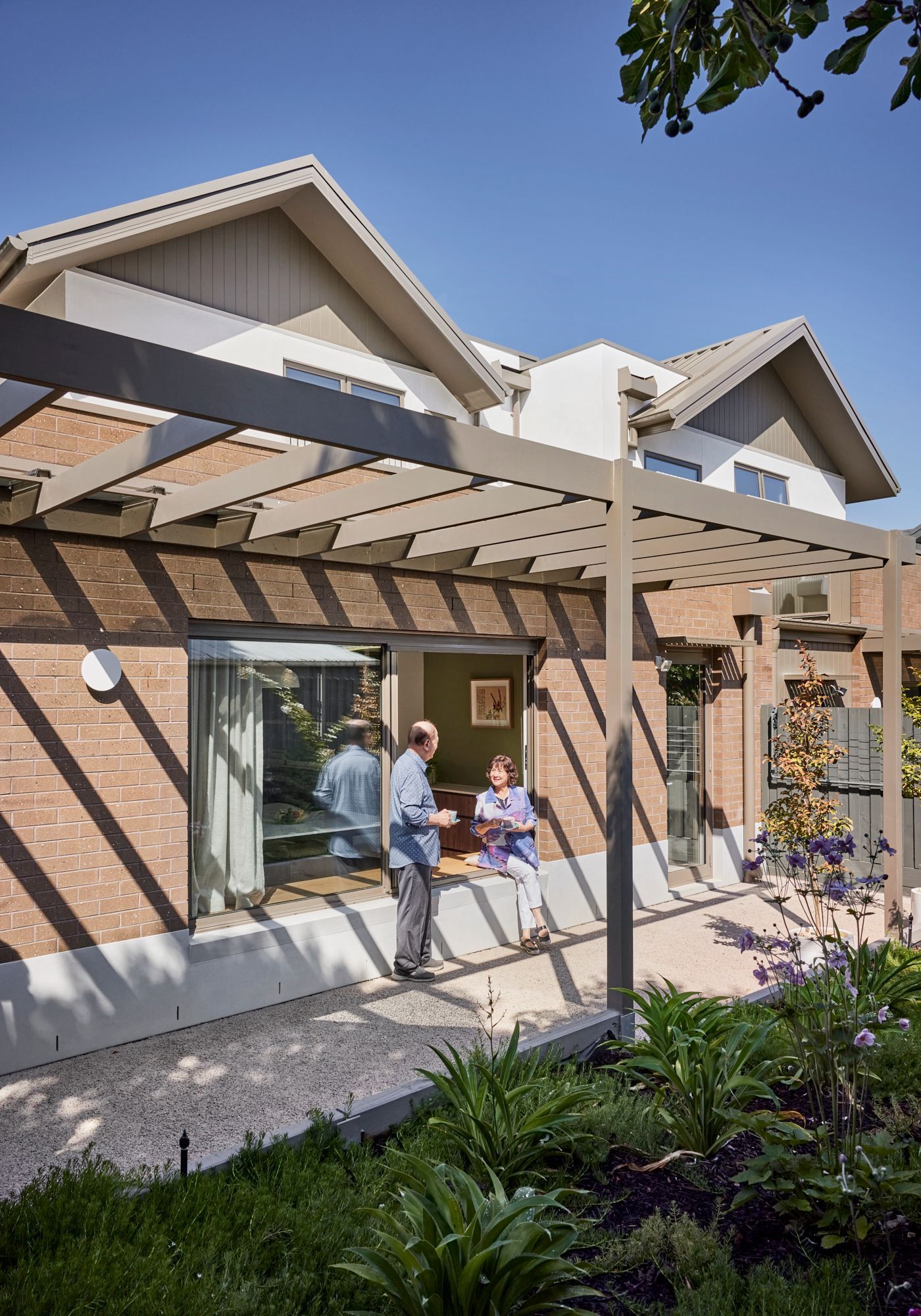 Two people are standing under a pergola in front of a house.