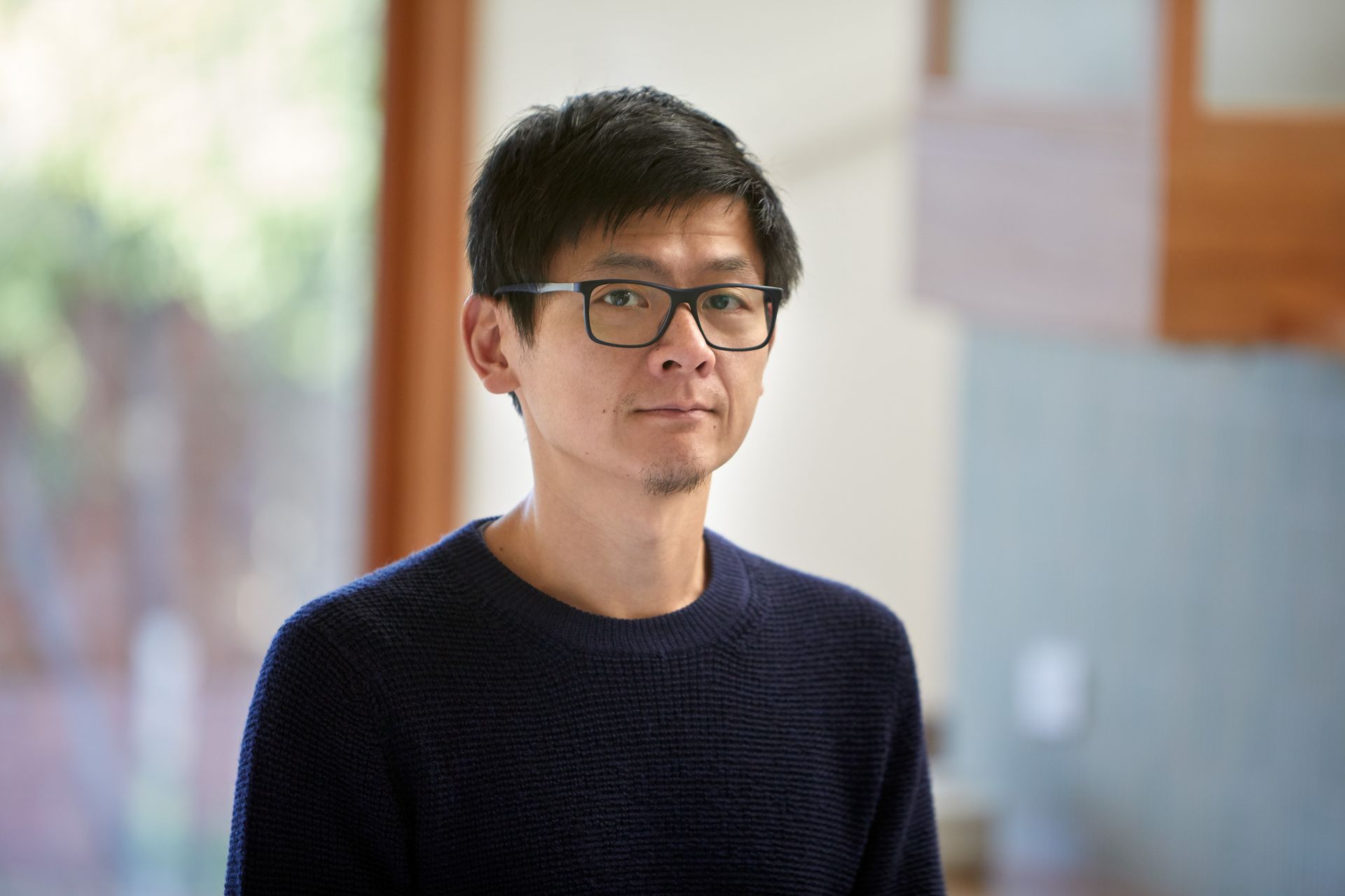 A man wearing glasses and a blue sweater is standing in front of a window.