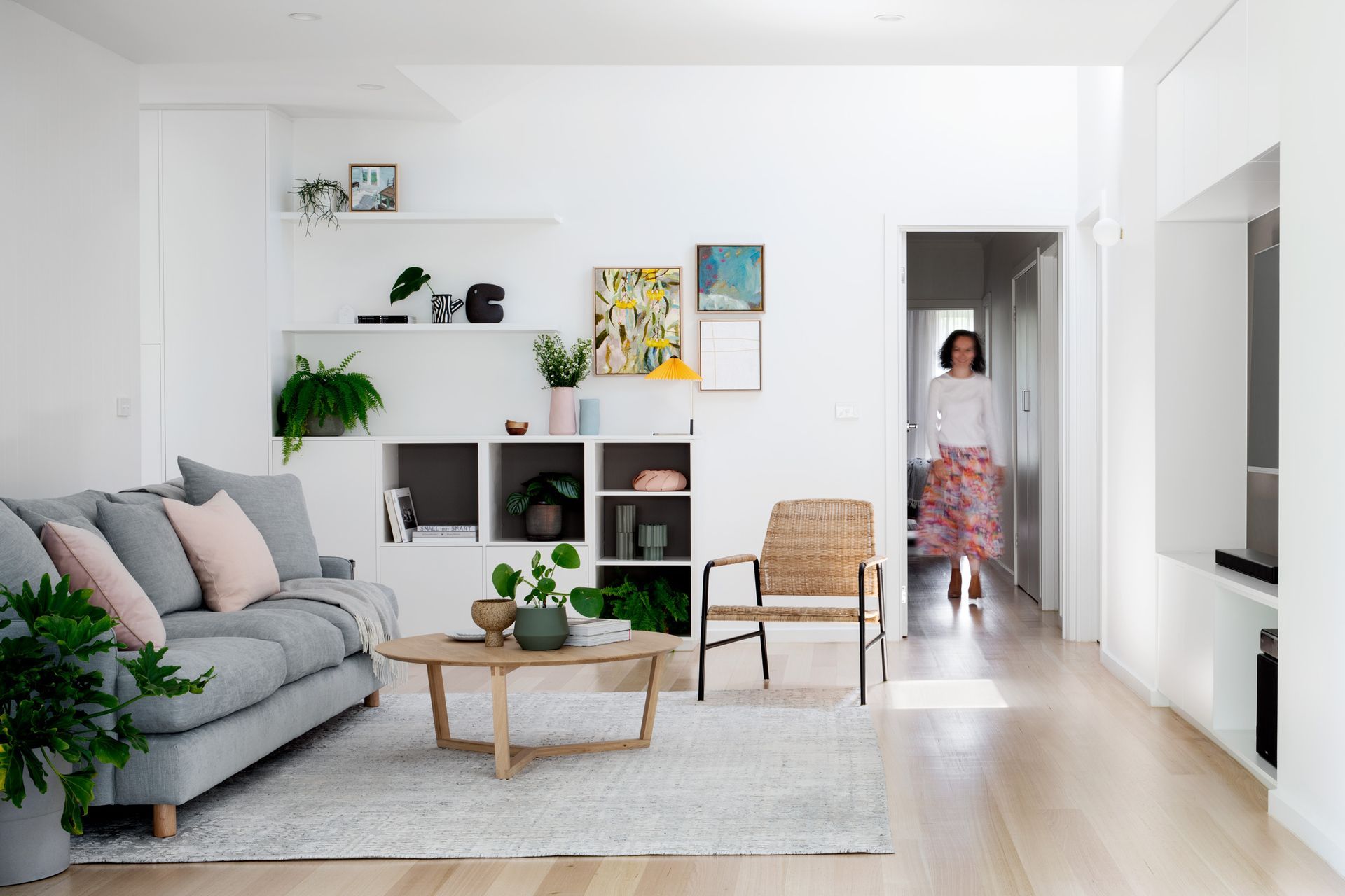 A woman is walking into a living room with a couch , chair , coffee table and shelves.