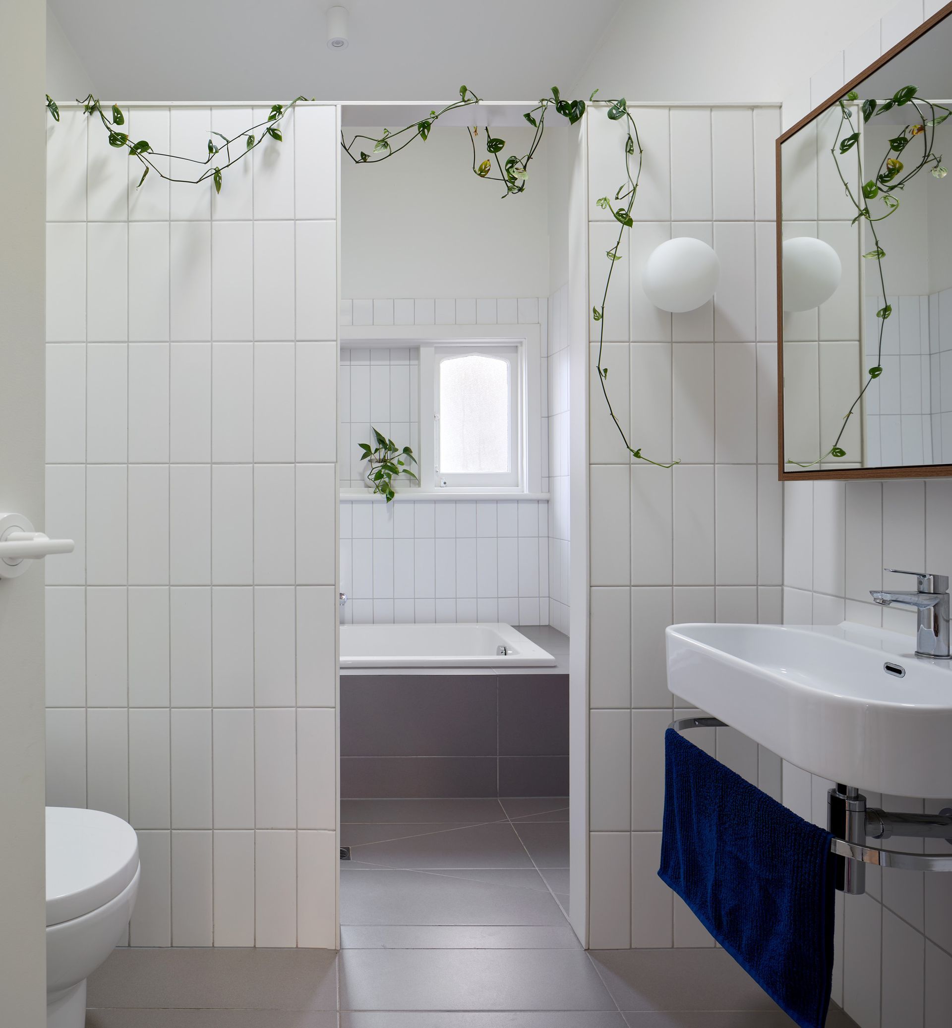 A modern bathroom with white tile walls, a toilet, sink, and bathtub. Green vines decorate the walls.