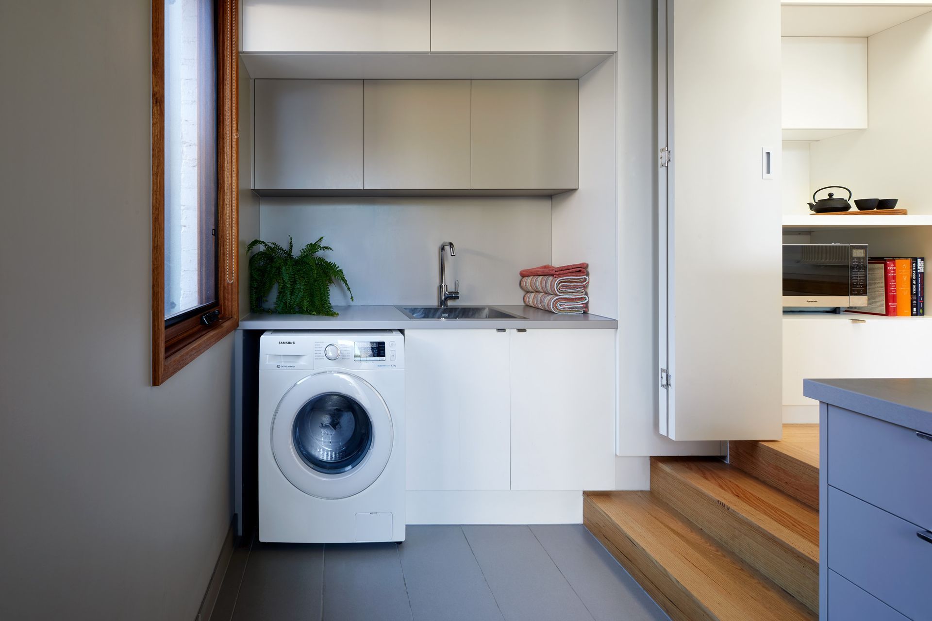 A small, modern laundry room with a washing machine, sink, and cabinets. Wooden steps lead to a separate area with a microwave.