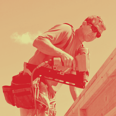 Man using a drywall screw gun on a wall, wearing a hard hat, glasses, and gloves.