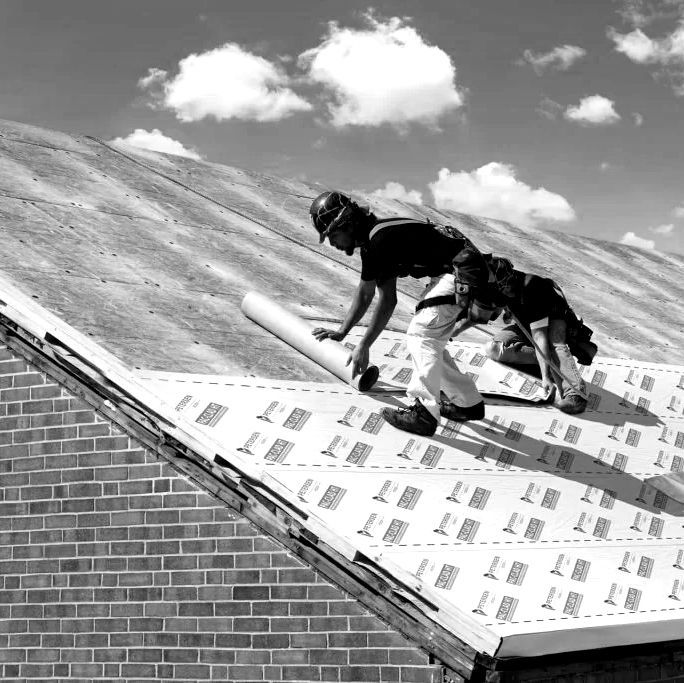 Roofer rolls out roofing material on a sloped roof. The sky is partly cloudy and the building has a brick facade.